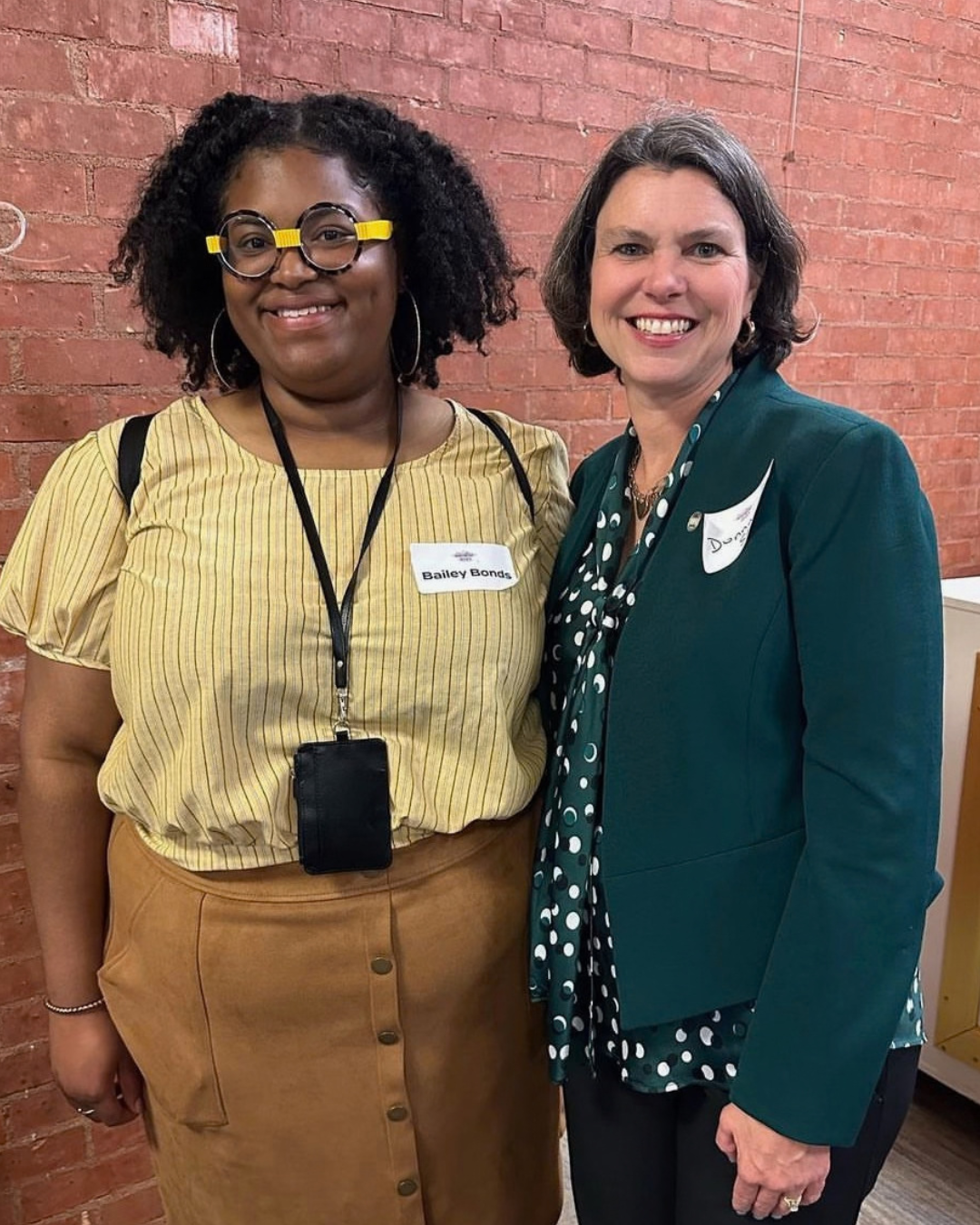 Two women smiling together at an event, one wearing yellow with round glasses, the other in a green blazer.