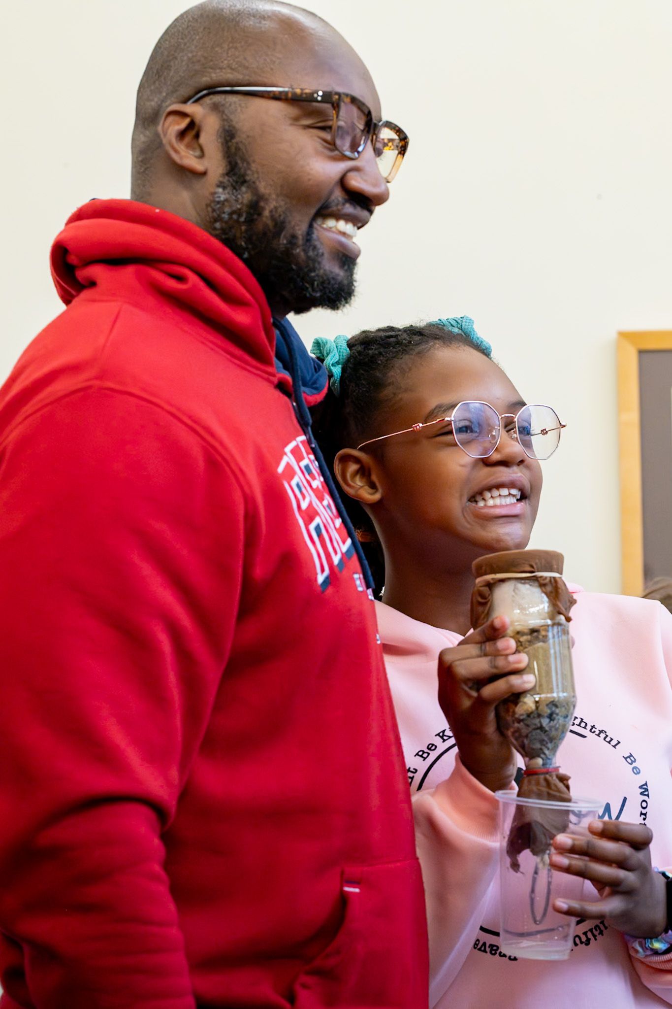 A man in a red hoodie and a young girl with blue hair smile while holding a glass jar together.