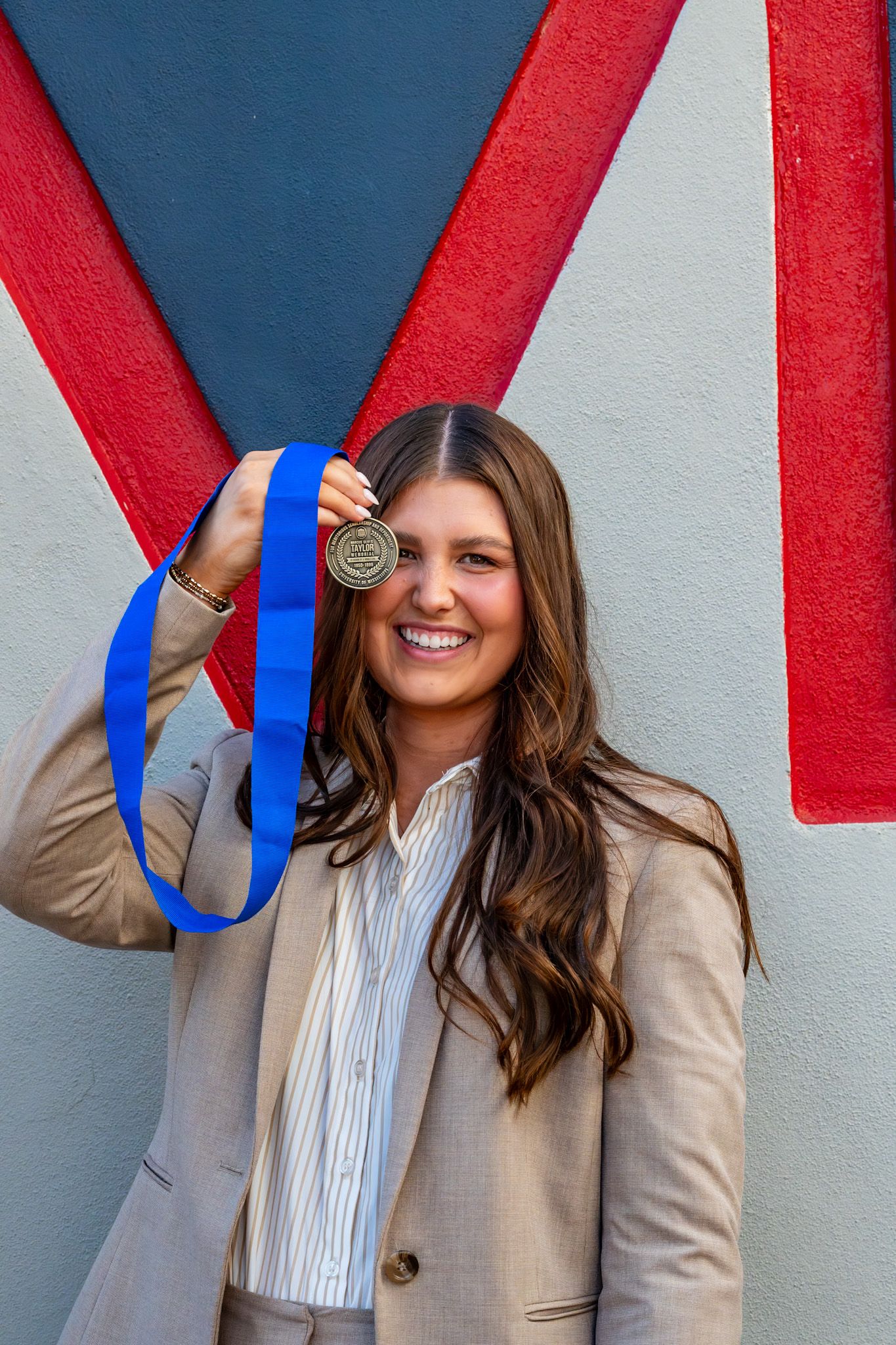 Smiling woman holding gold medal with blue ribbon near face outdoors.