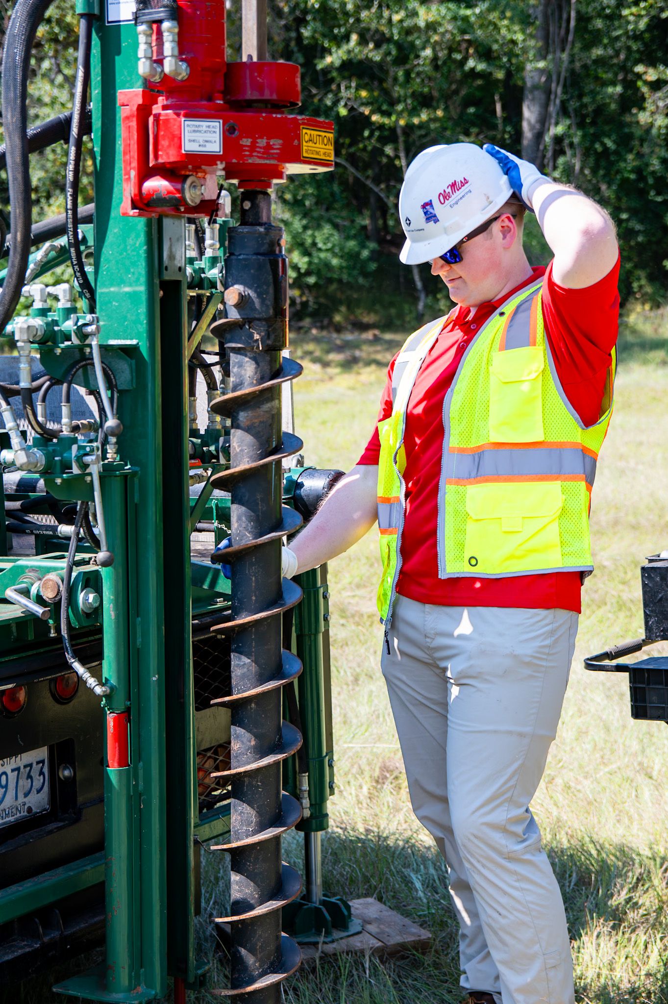 Construction worker in safety vest and hard hat operating a drilling machine on a green field job site.