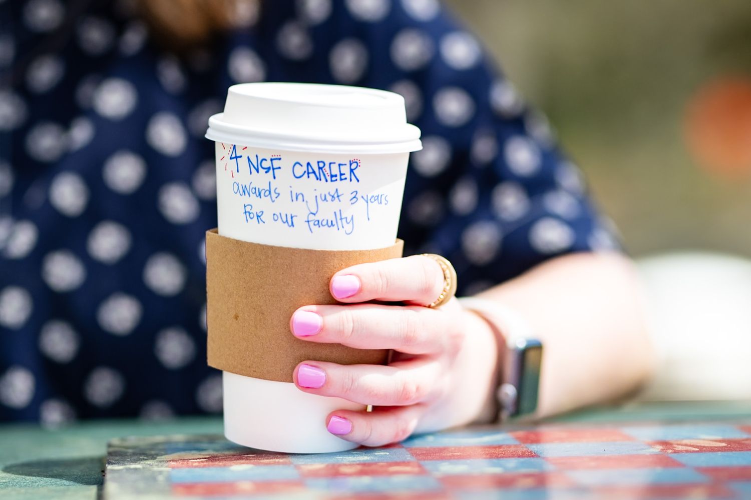 Woman holding a disposable coffee cup with a cardboard sleeve and handwritten career message, wearing pink nail polish.