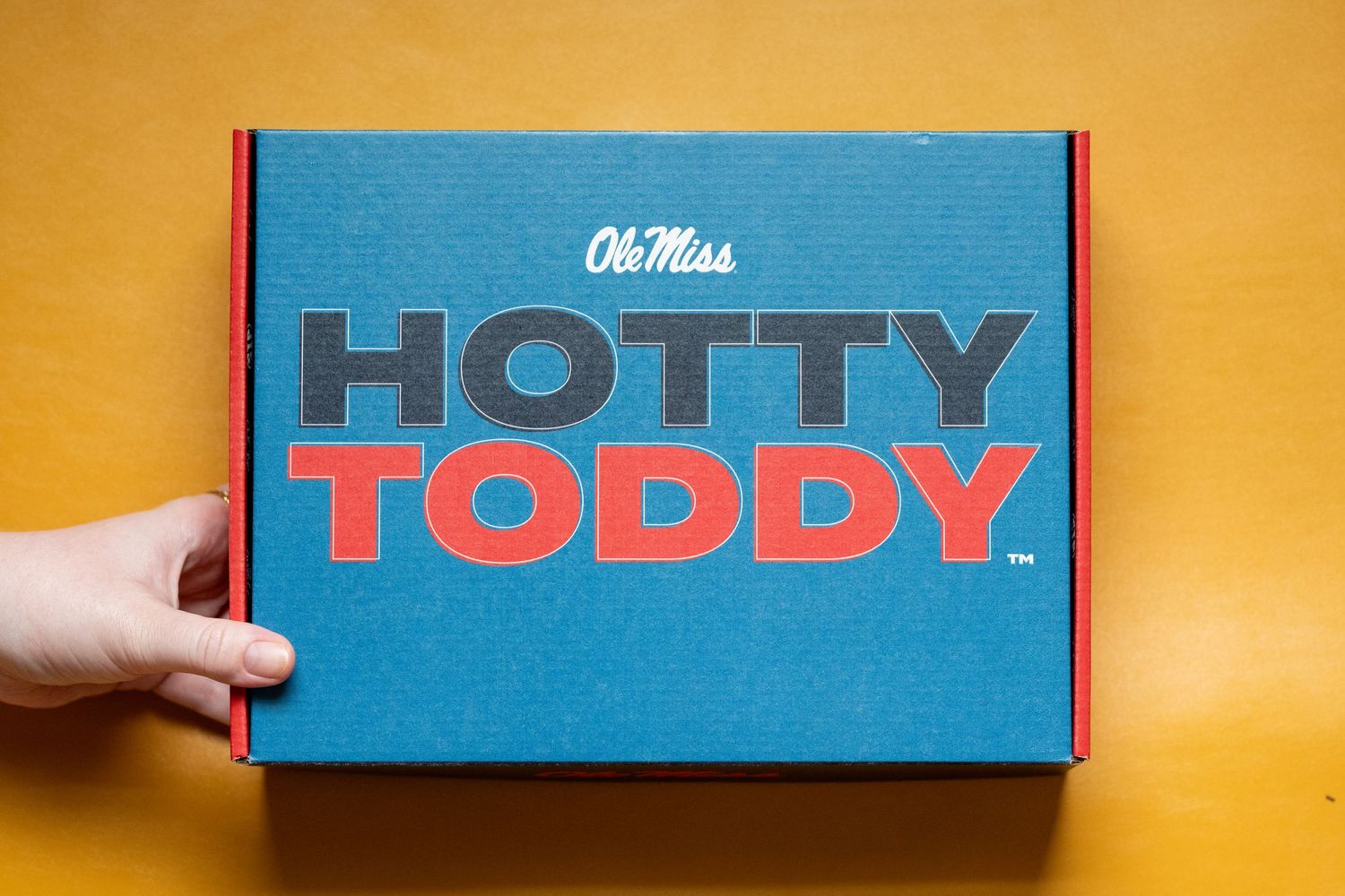Hand holding a teal Ole Miss Hotty Toddy branded gift box with bold red and black typography on yellow background.
