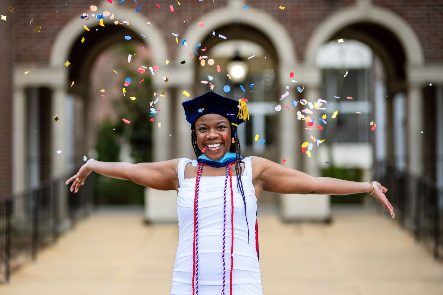 A graduate celebrates with outstretched arms as confetti falls during commencement at an ornate stone archway.