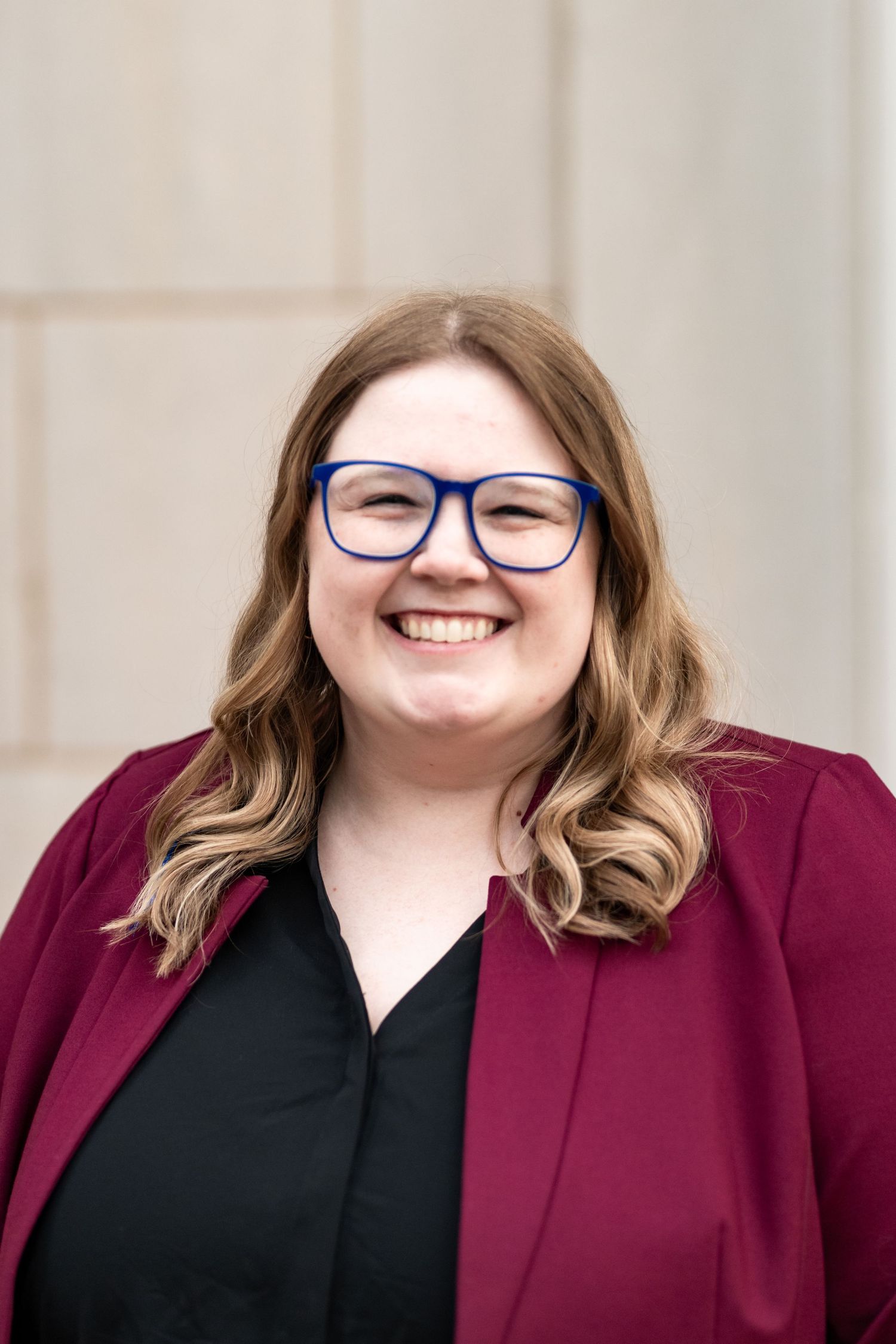 Professional headshot of someone in a burgundy blazer and black top smiling warmly while wearing blue-framed glasses.