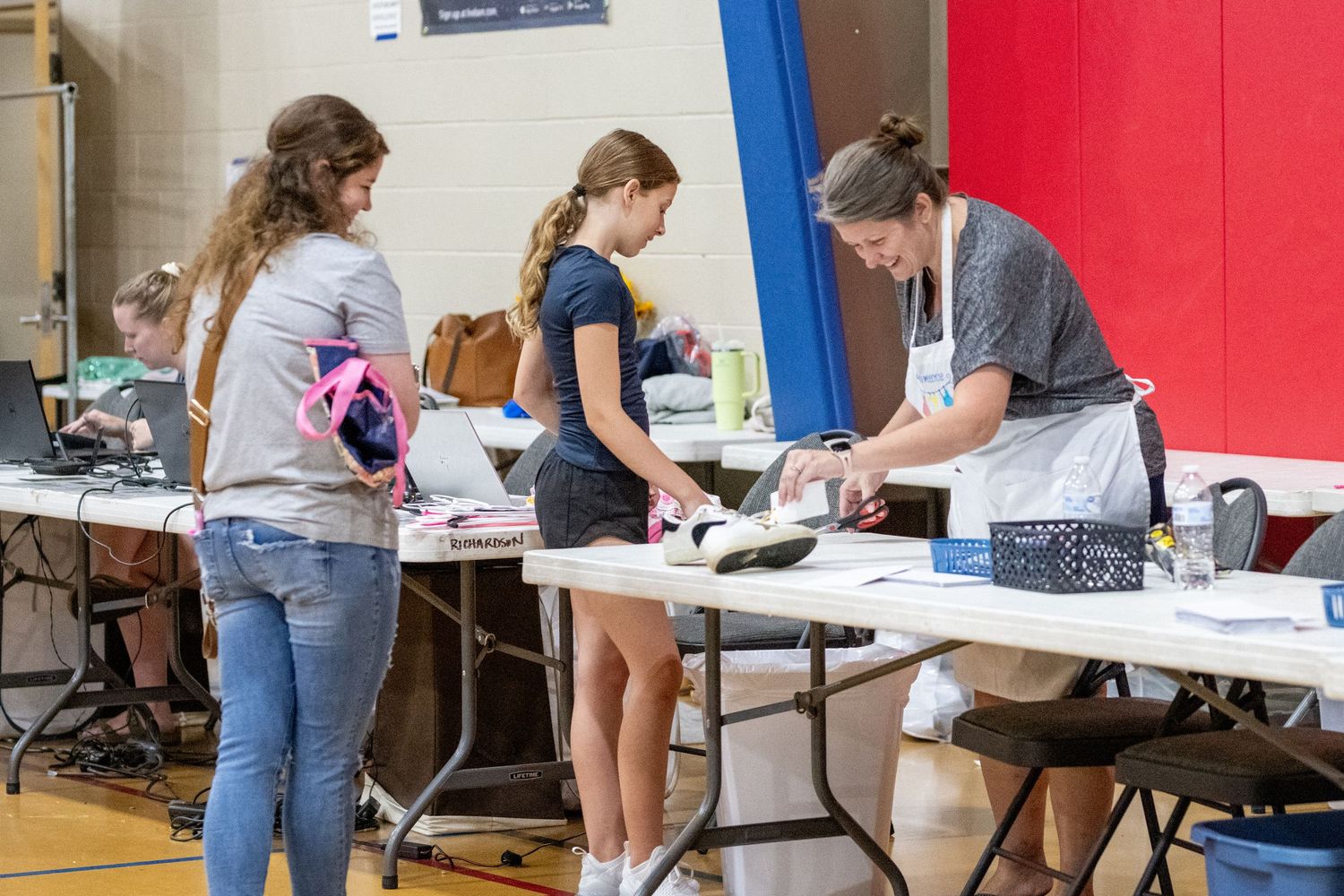 Students register at a school event table in a gymnasium, with a woman assisting two girls with paperwork.