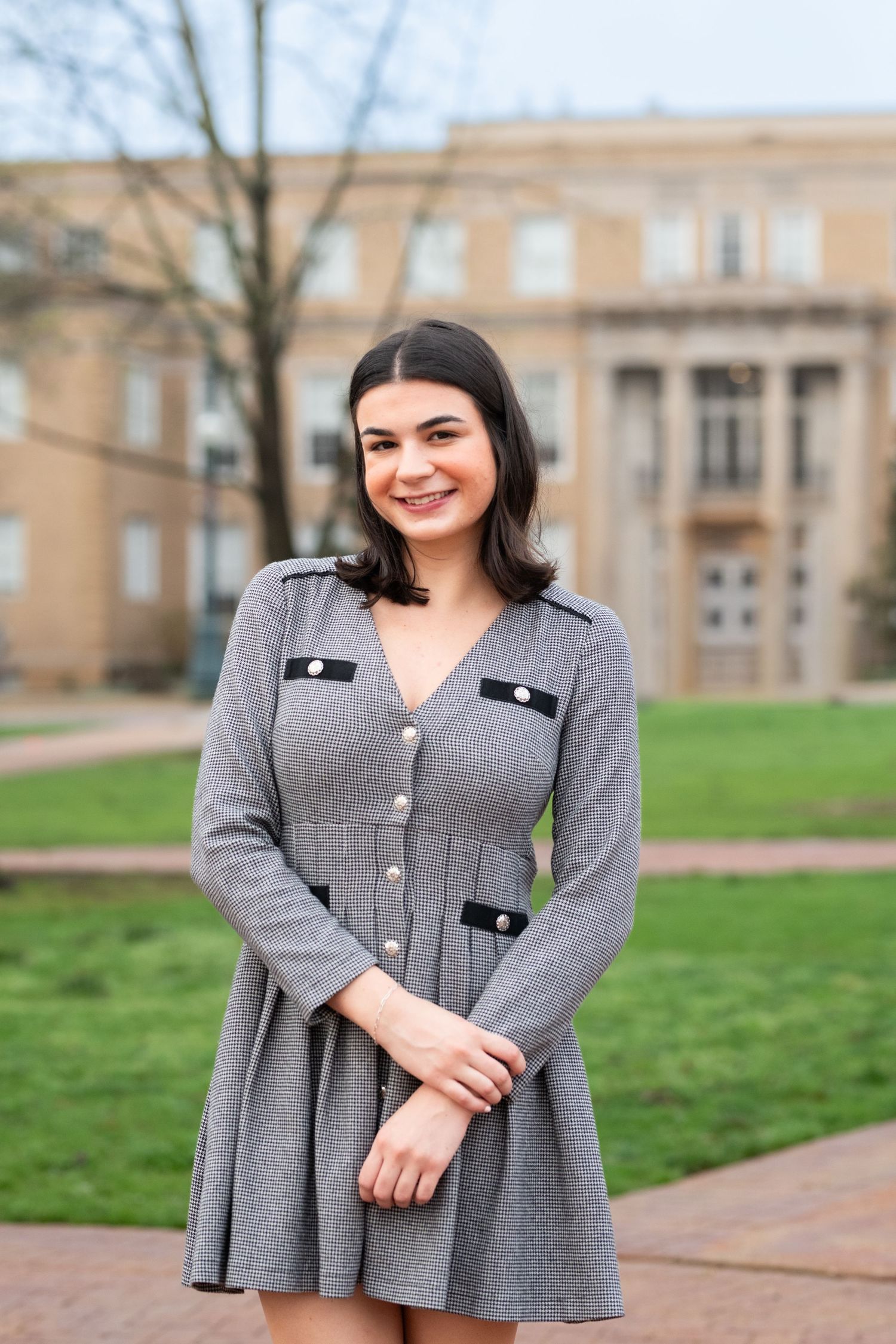 A woman in a gray button-up peplum jacket poses outdoors in front of a tan building on a grassy lawn.