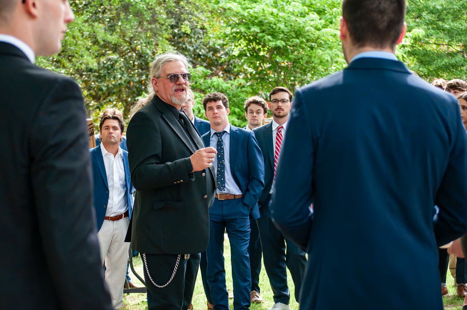Older man in black blazer with sunglasses speaking to group of young men in suits outdoors.