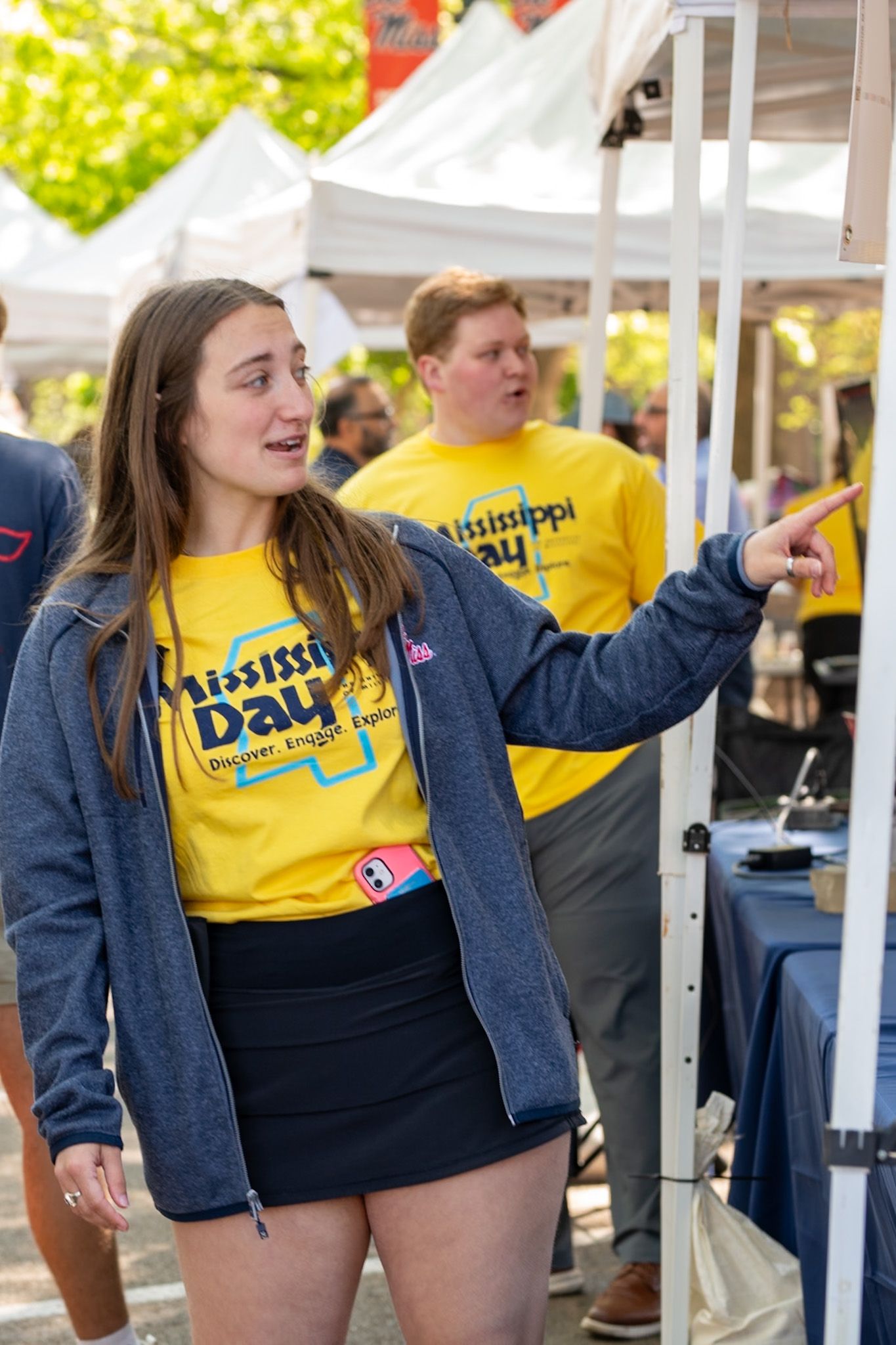 Young woman in Mississippi Day yellow shirt pointing at outdoor event with another volunteer in background.