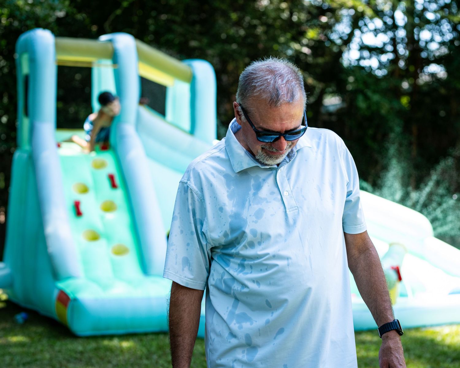 Older man in sunglasses and wet shirt stands near inflatable bounce house slide at outdoor party.