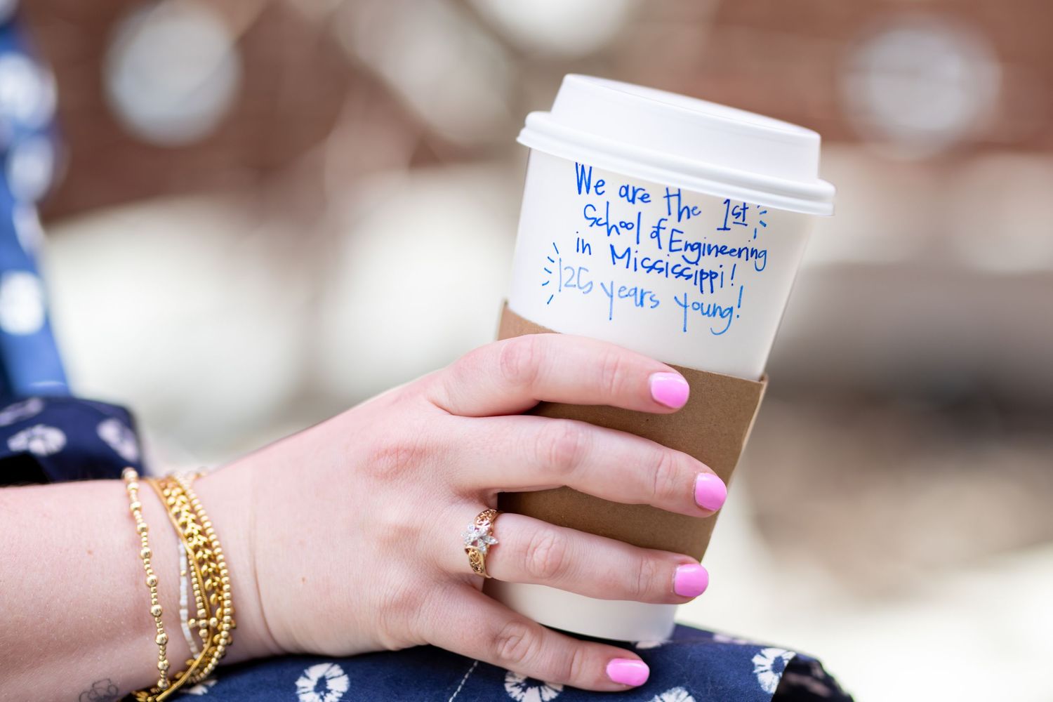 Hand holding a coffee cup with handwritten text reading We are the 1st School of Engineering in Mississippi, pink nails and gold bracelets.