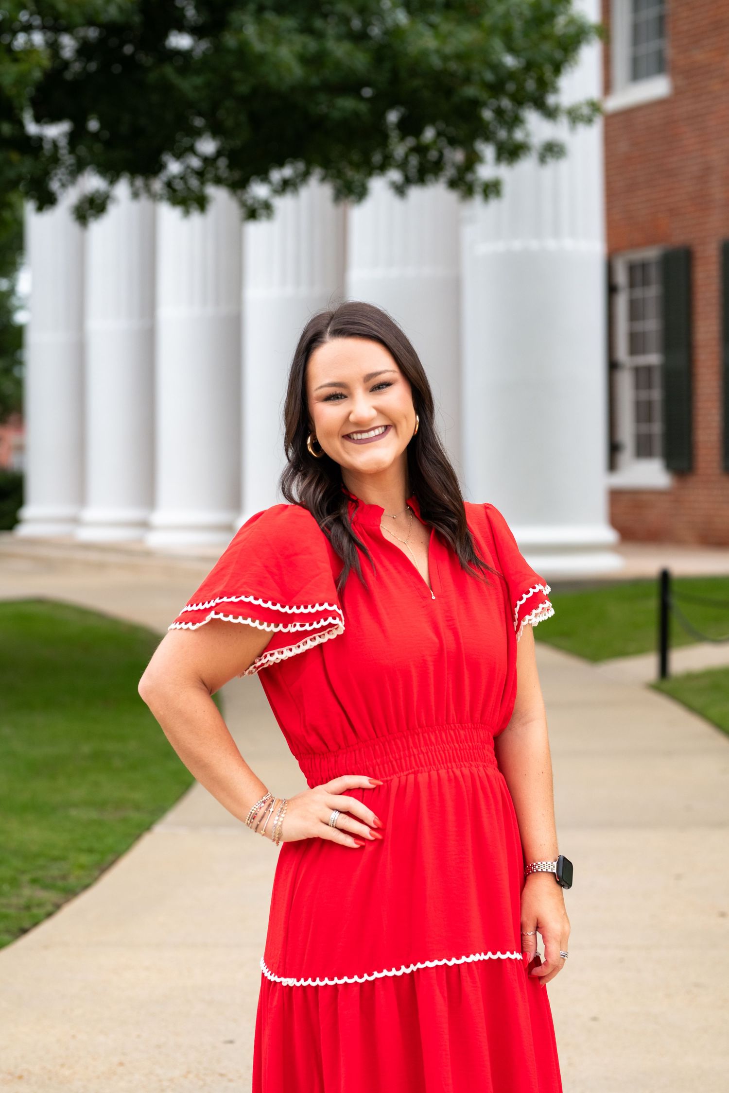 Smiling woman in red dress with white trim posing outdoors near white columns of a brick building.