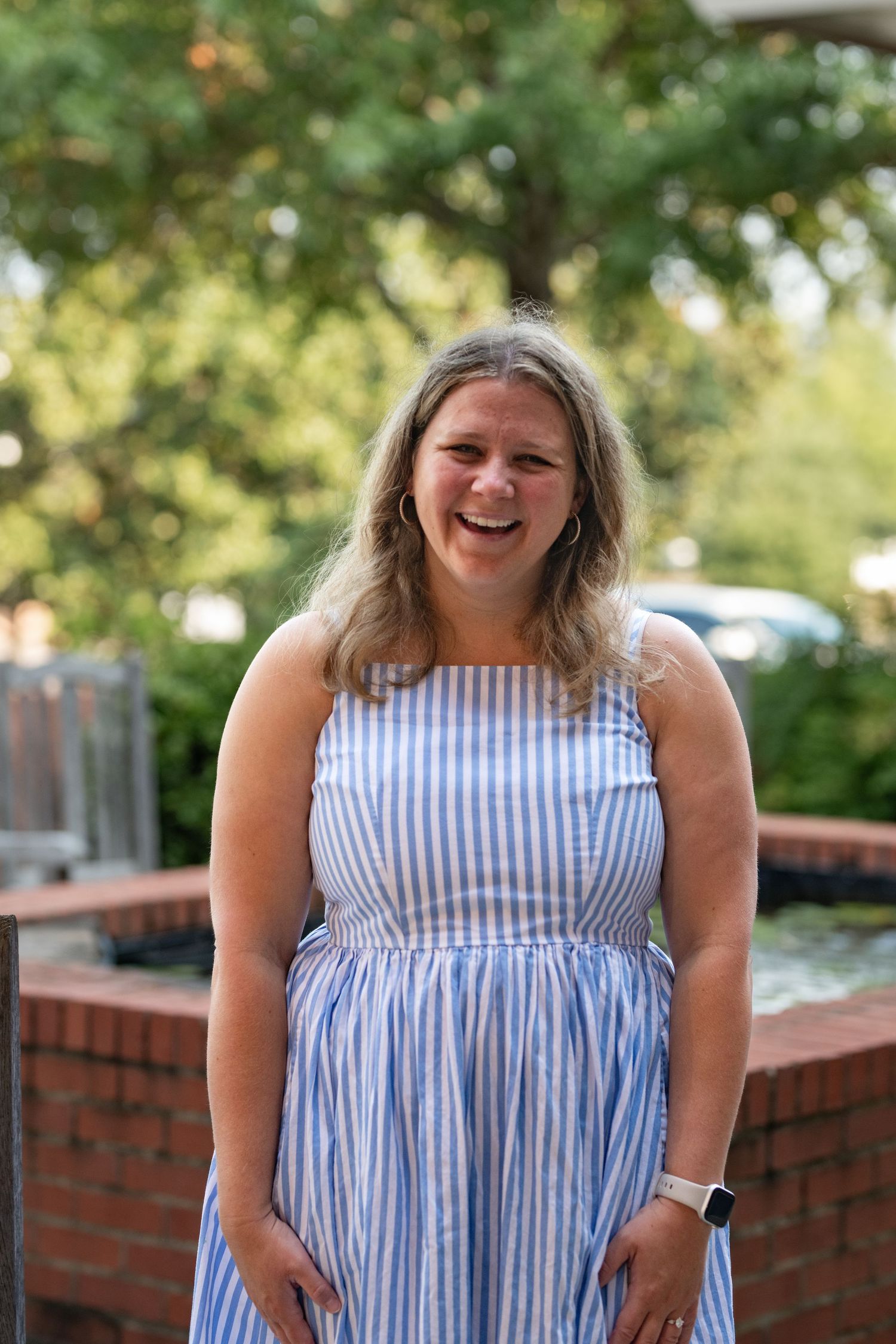 Smiling woman in blue striped sundress standing outdoors near brick fountain with green trees in background.
