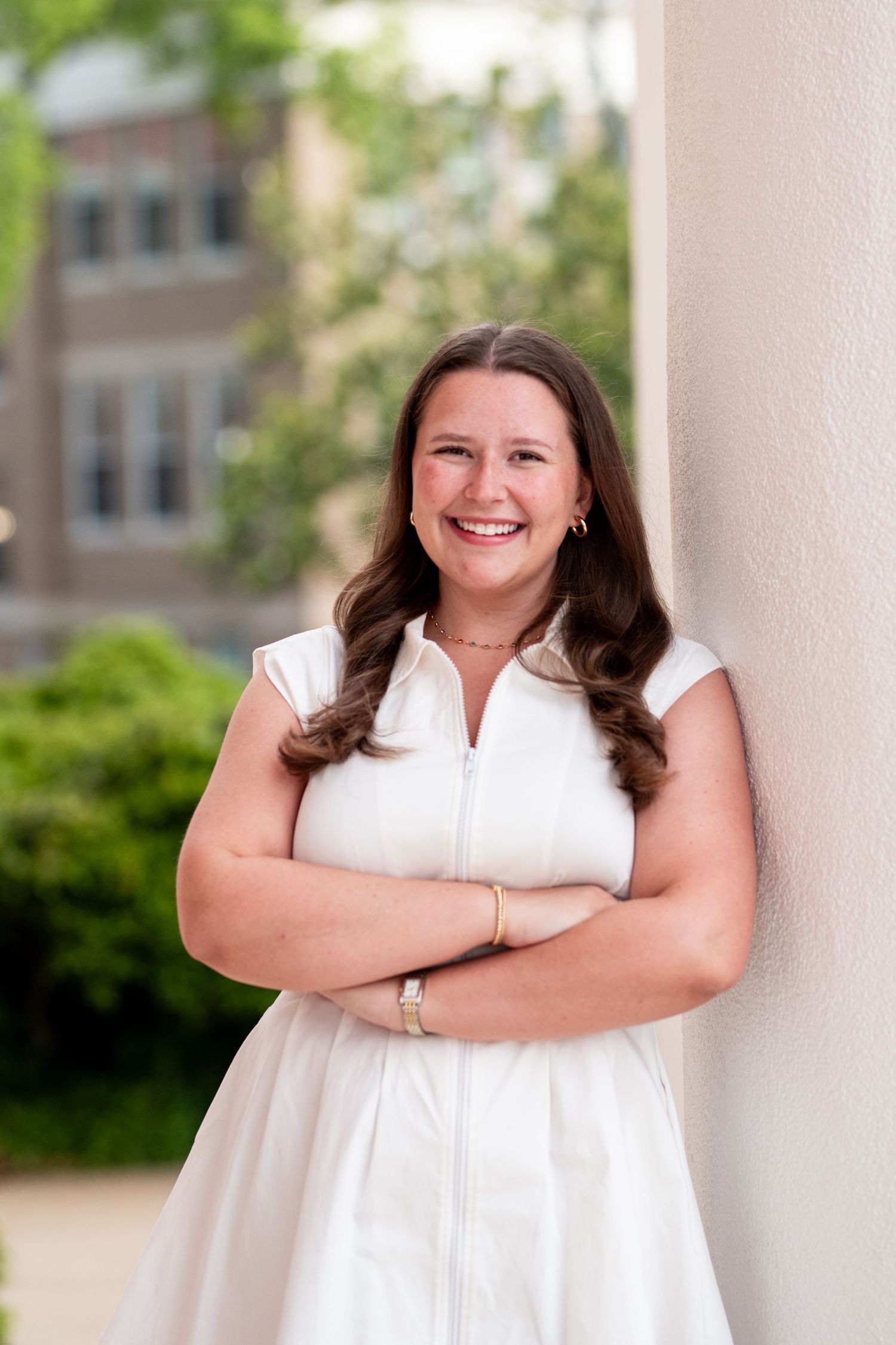 Smiling young woman in white sleeveless dress leaning against column with arms crossed outdoors on college campus.