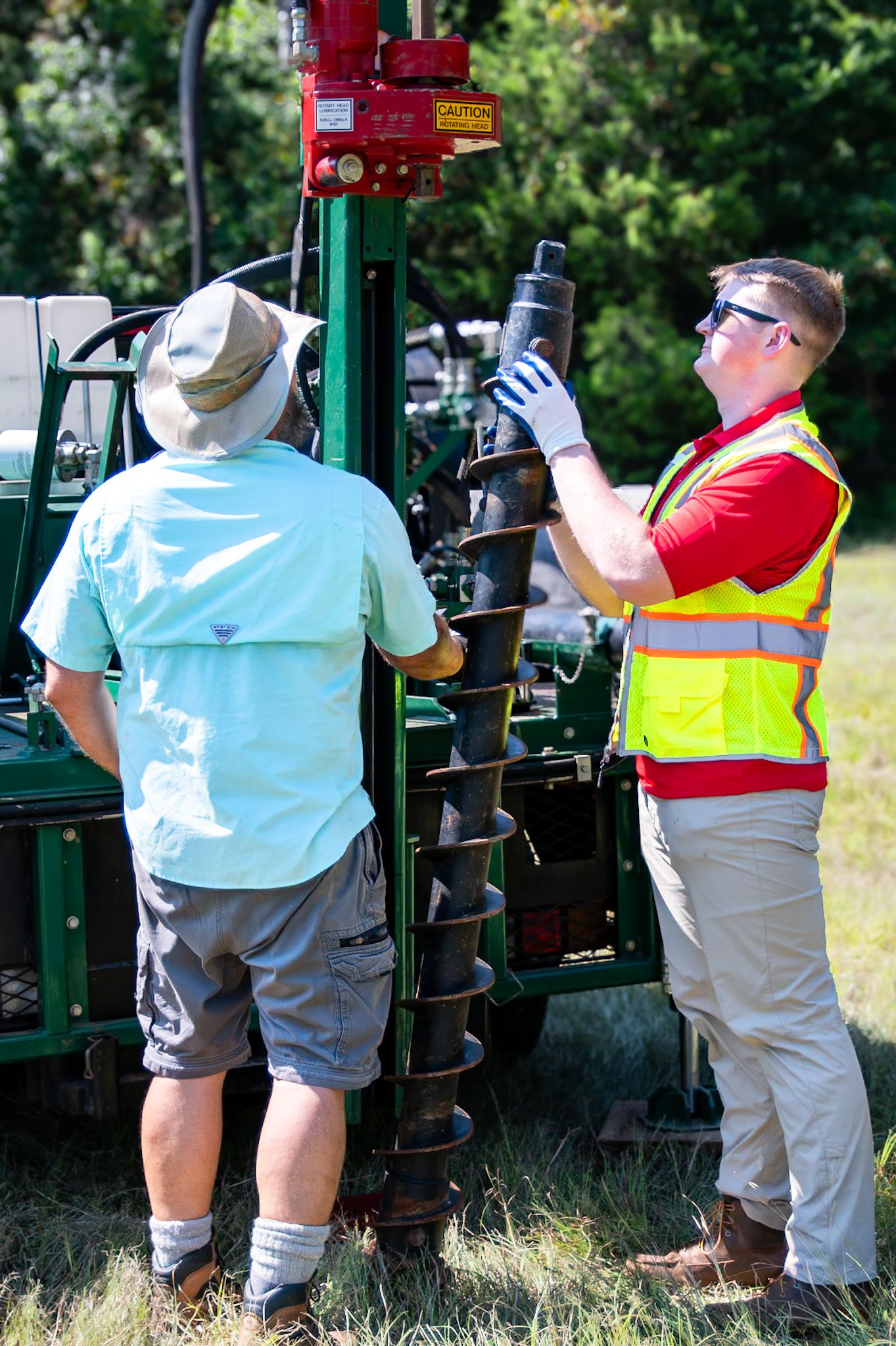 Two workers in safety gear operating a ground drilling machine outdoors on a sunny day.