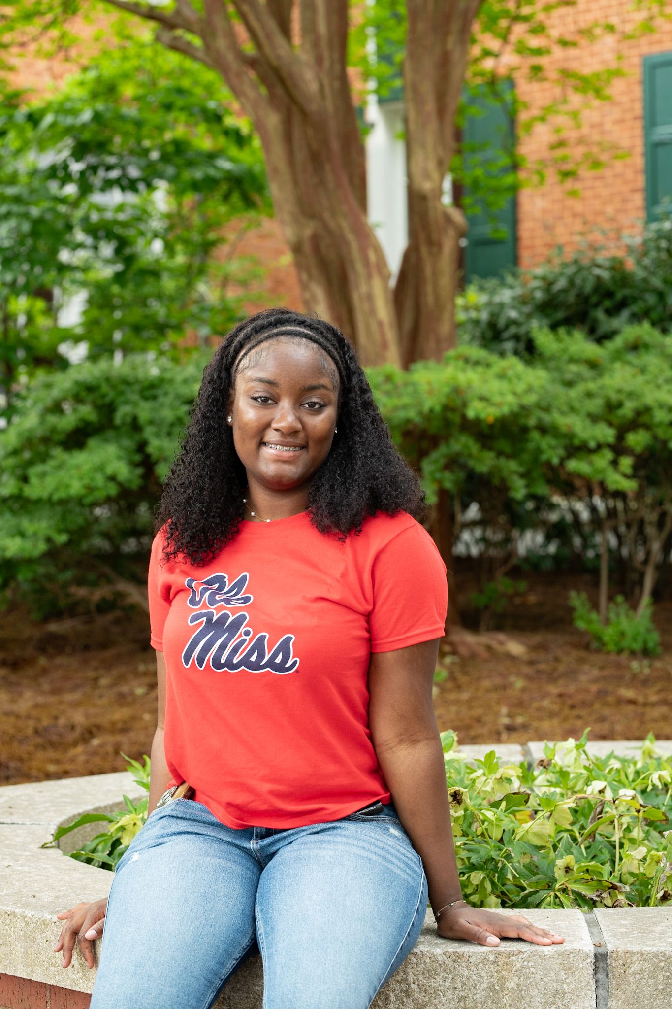 Young woman in Ole Miss red t-shirt sitting on stone wall surrounded by lush green campus landscaping.