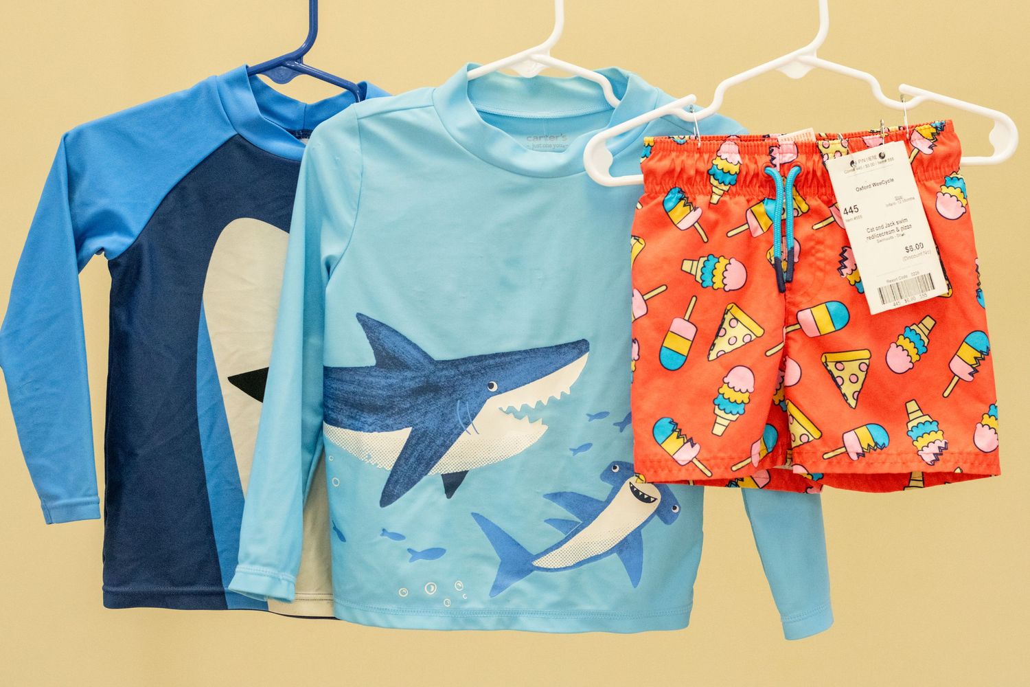 Three boys' swimwear pieces: two shark-print rash guards in blue and red patterned swim trunks on hangers.