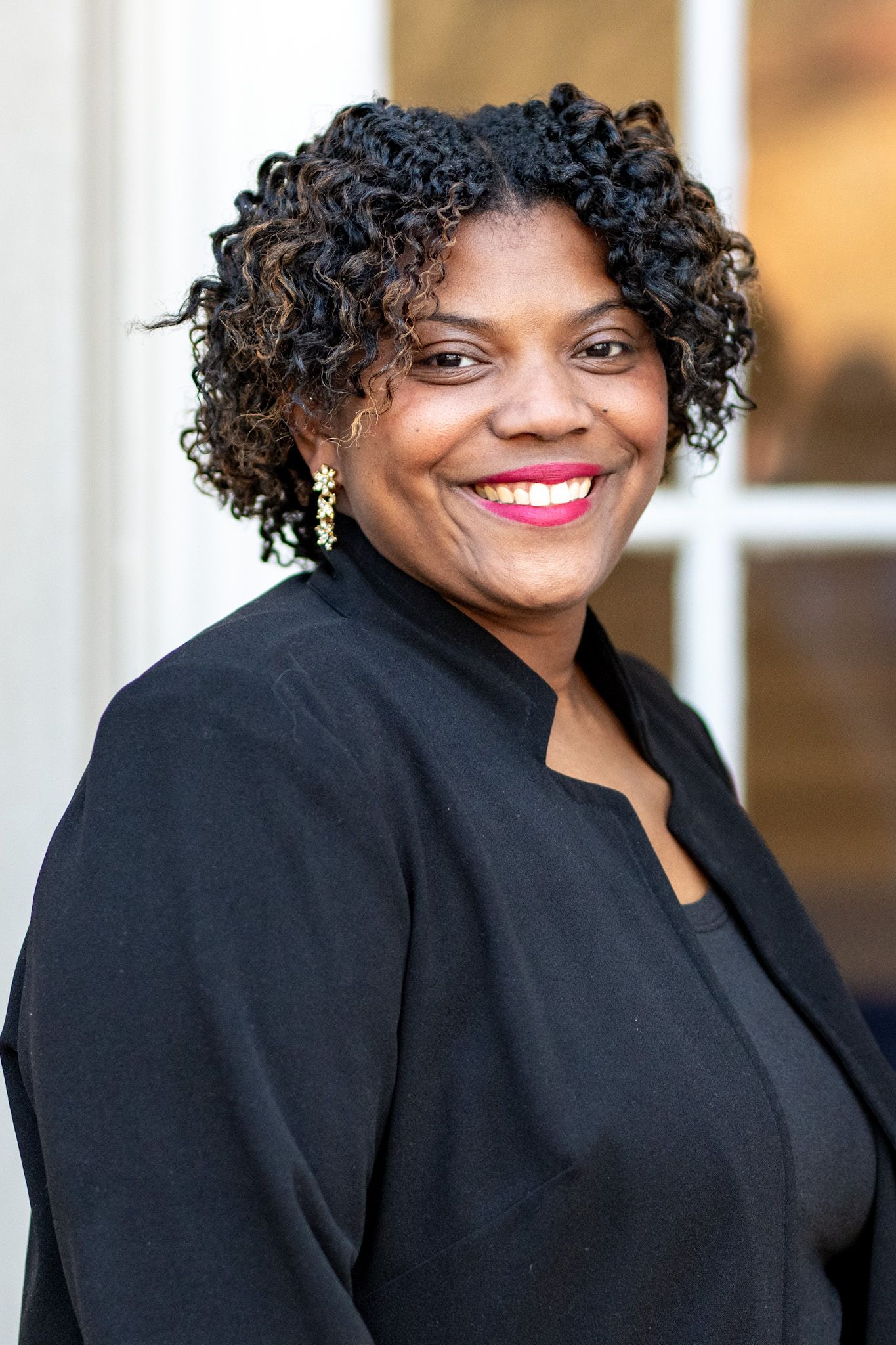 Professional headshot of someone in a black blazer with natural curly hair and bright lipstick smiling warmly at camera.