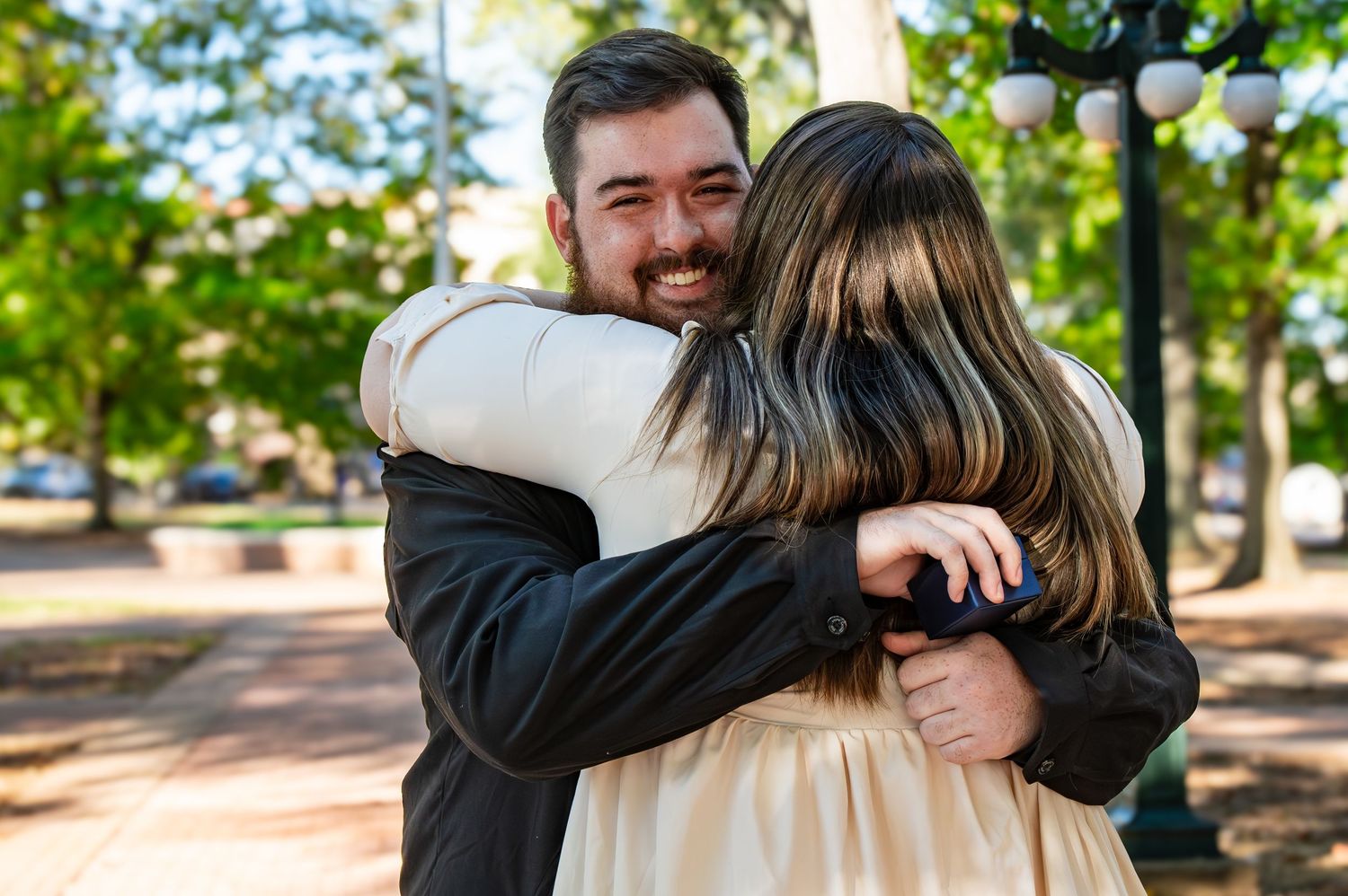 A couple shares a warm embrace on a tree-lined path in the park on a sunny afternoon.
