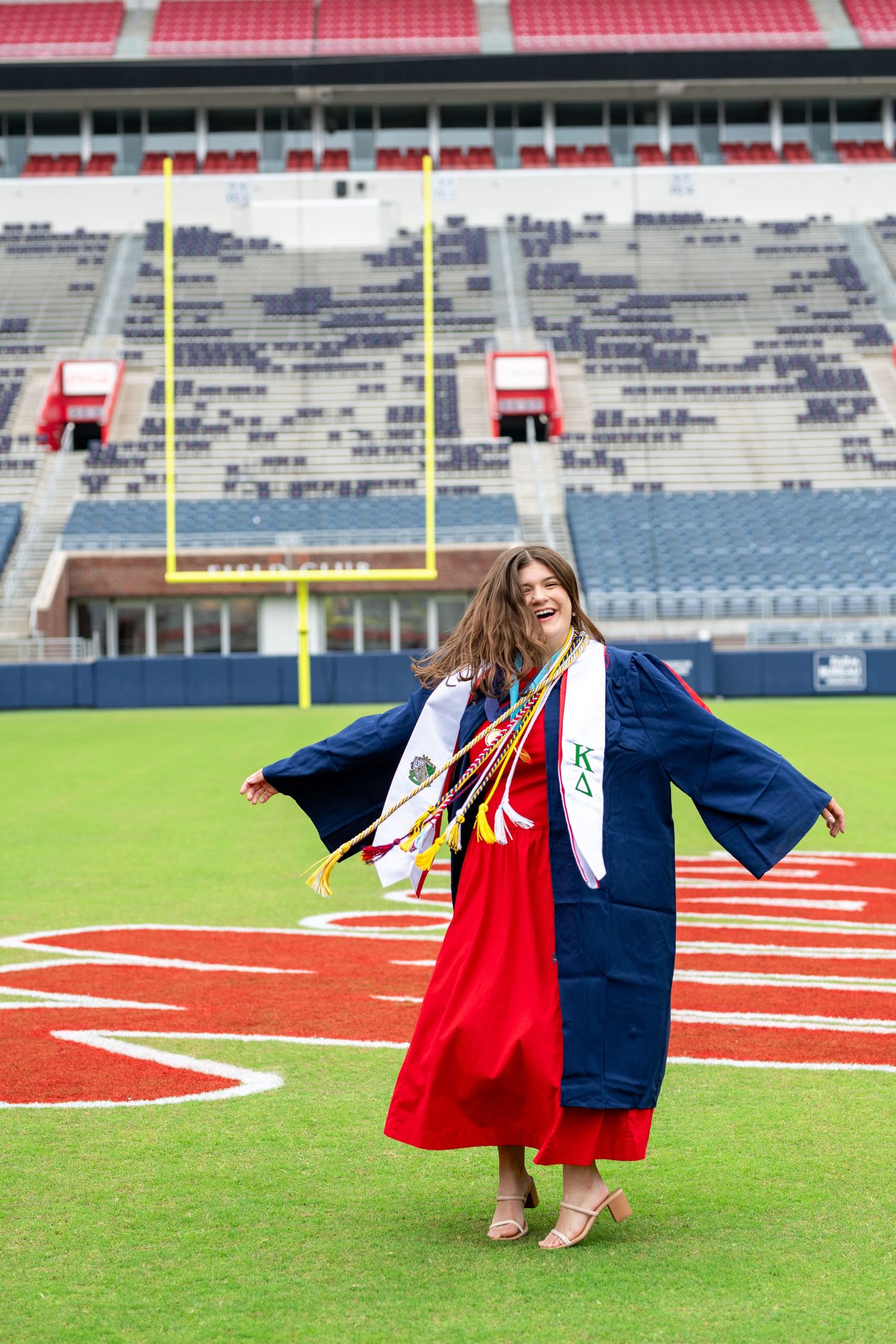 Graduate in red dress and navy blue gown posing on football field with stadium seats in background.