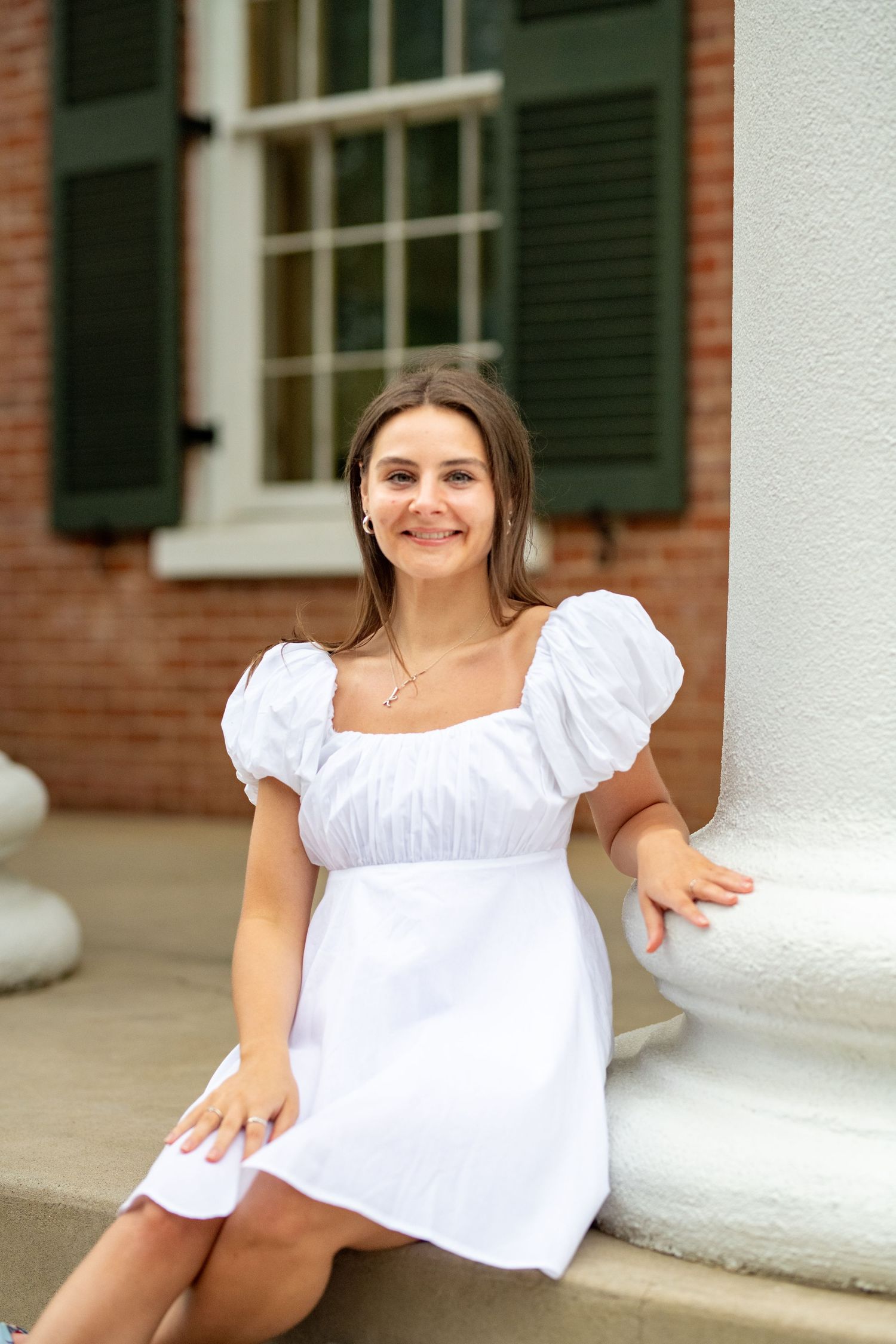 A person in a white dress poses on a classical brick building porch with white columns.