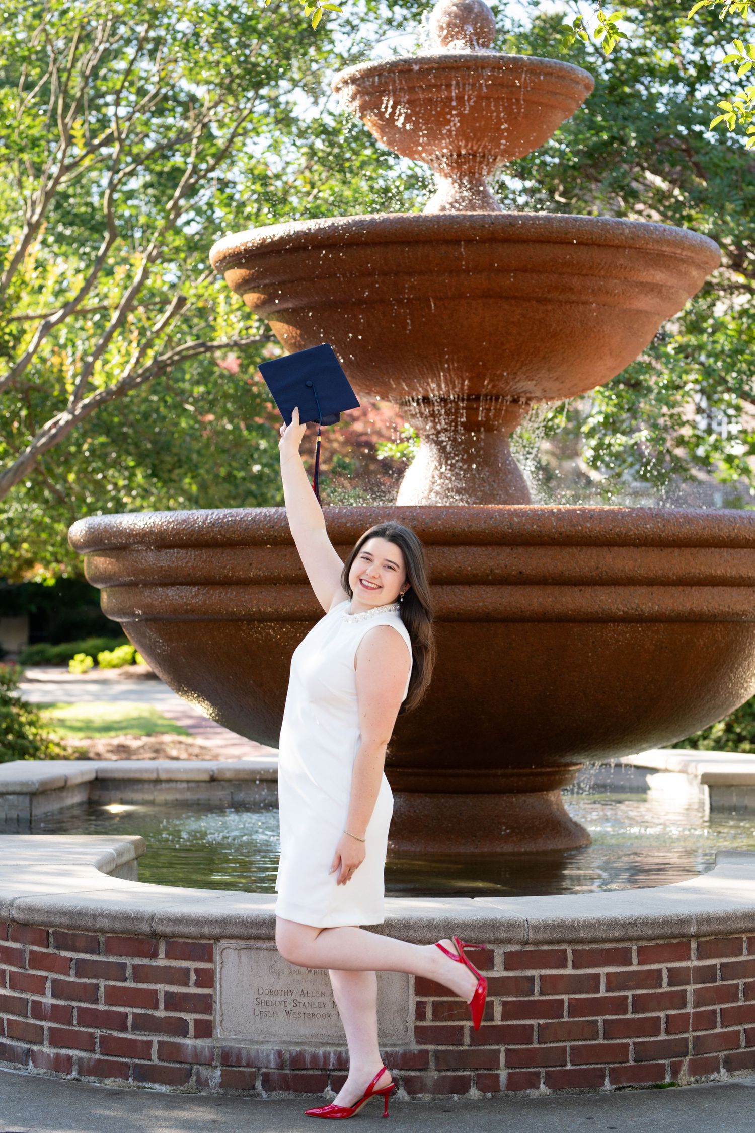 Young woman in white dress and red heels celebrates graduation by fountain, holding diploma aloft with one leg raised.