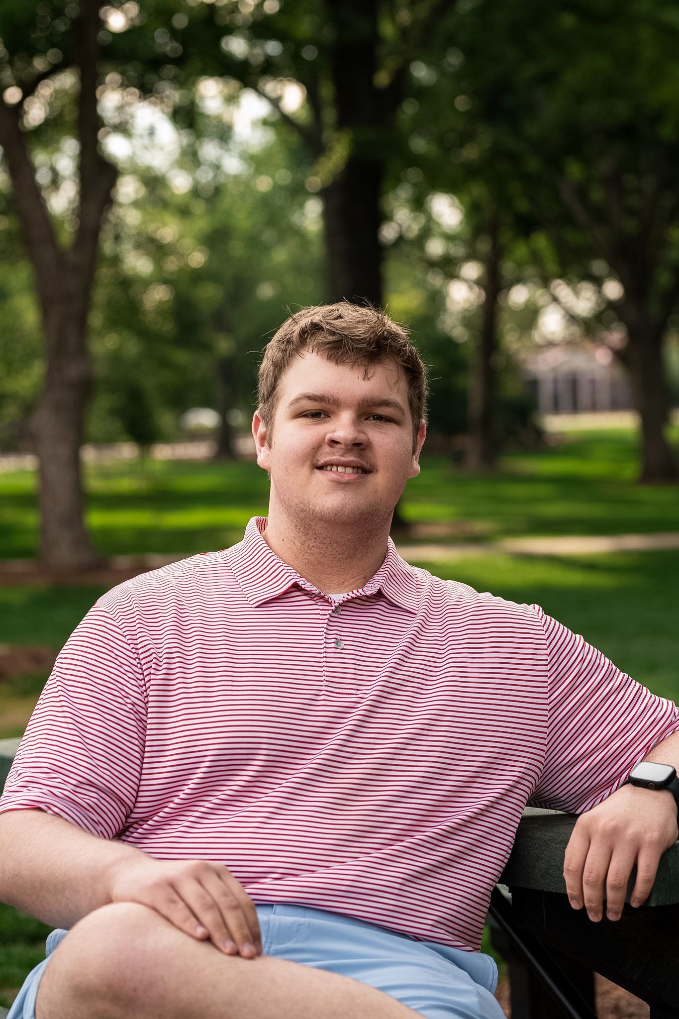Young man in pink striped polo shirt smiling while sitting on bench in lush green park setting.