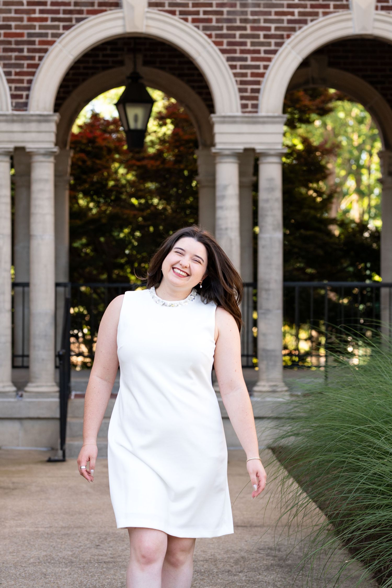 Smiling woman in white sleeveless dress walking near brick arches and stone columns outdoors.