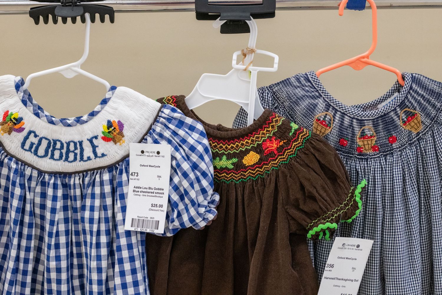 Three smocked girls dresses on hangers, including a blue gingham dress with Gobble embroidery for Thanksgiving.