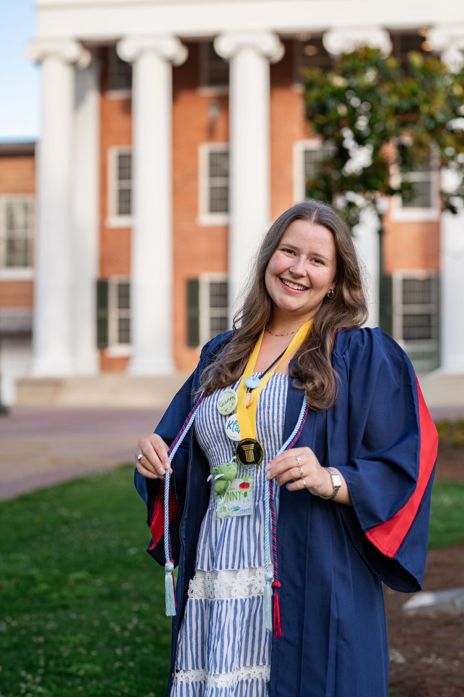 Smiling young woman in blue graduation gown holding cap outside brick university building with white columns.