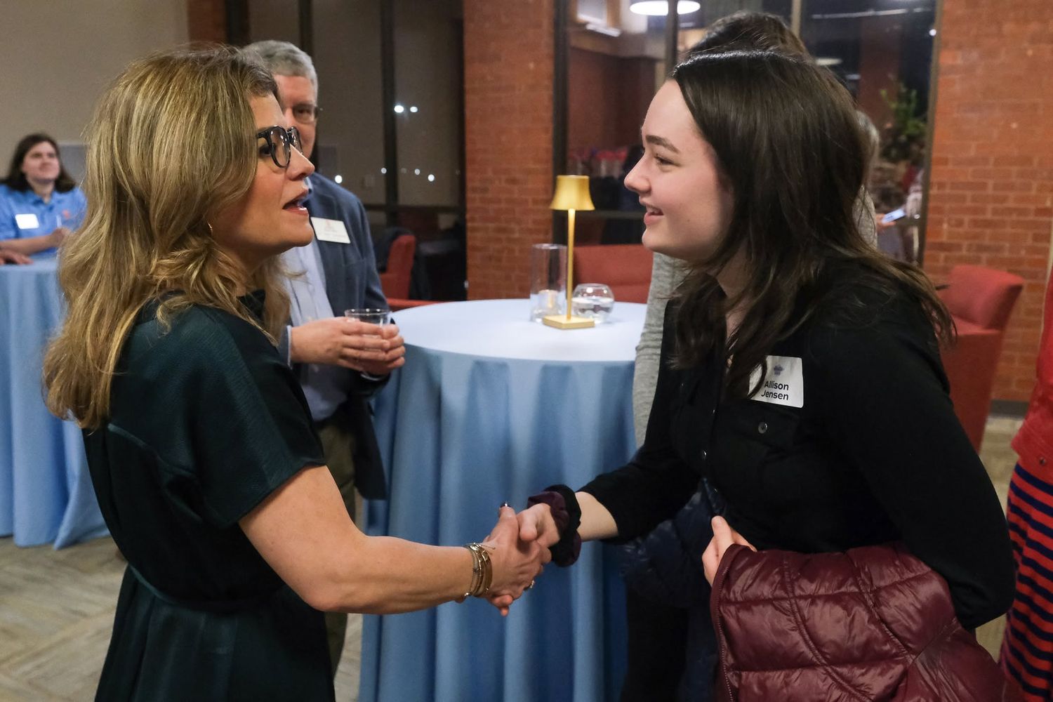 Two women shaking hands and smiling at a professional networking event with people gathered in the background.