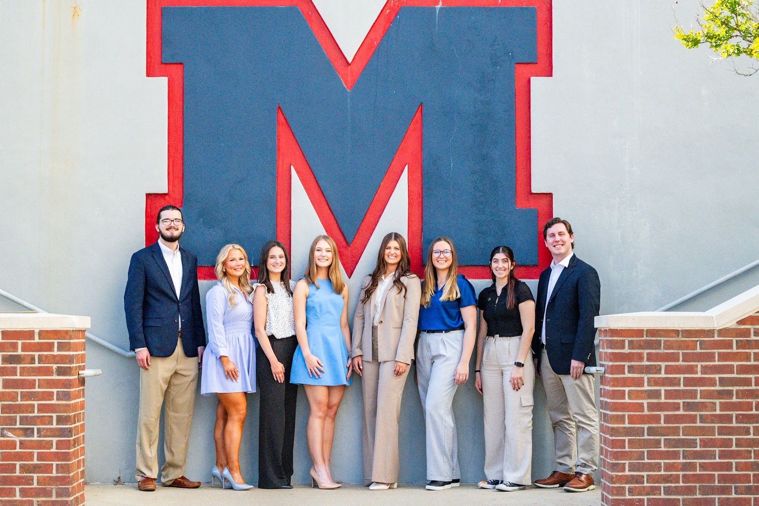 Group of eight young professionals posing in front of a large letter M logo on a brick wall outdoors.