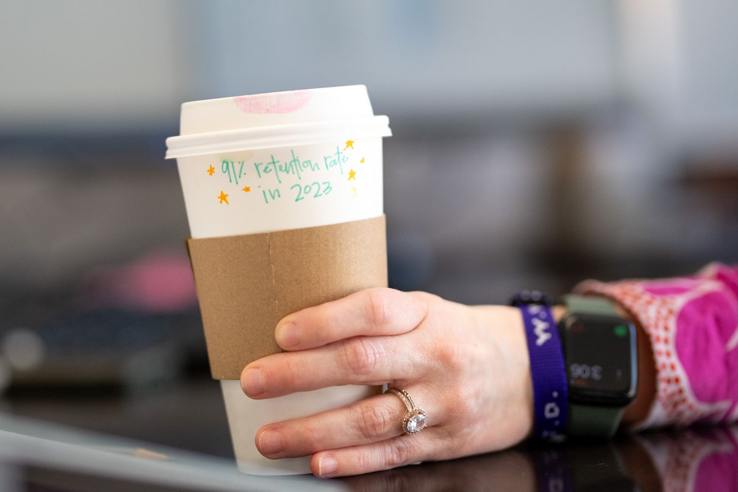 A hand wearing rings and a smartwatch holds a kraft paper coffee cup with colorful handwritten text.