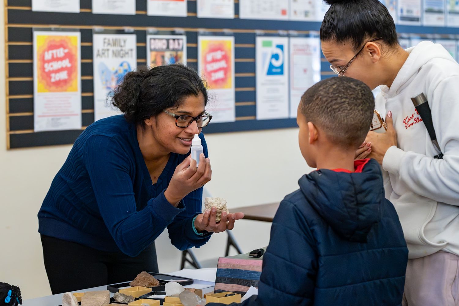 Teacher engages students in hands-on learning activity at classroom table during family art night event.