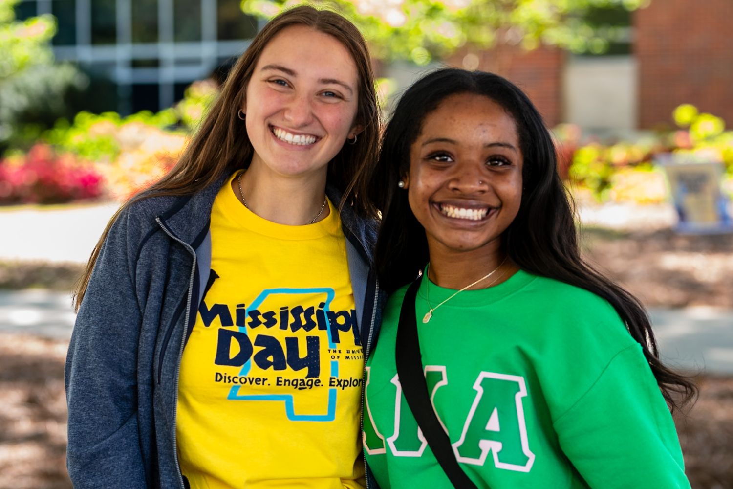 Two smiling students in college apparel, one wearing a yellow 'Mission Day' shirt and one in a green Greek letter shirt.