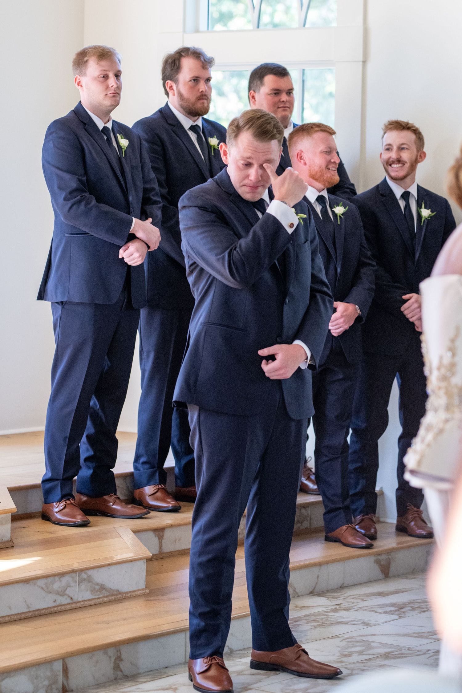 Emotional groomsman wipes tears during wedding ceremony while standing with other groomsmen in navy suits and brown shoes.