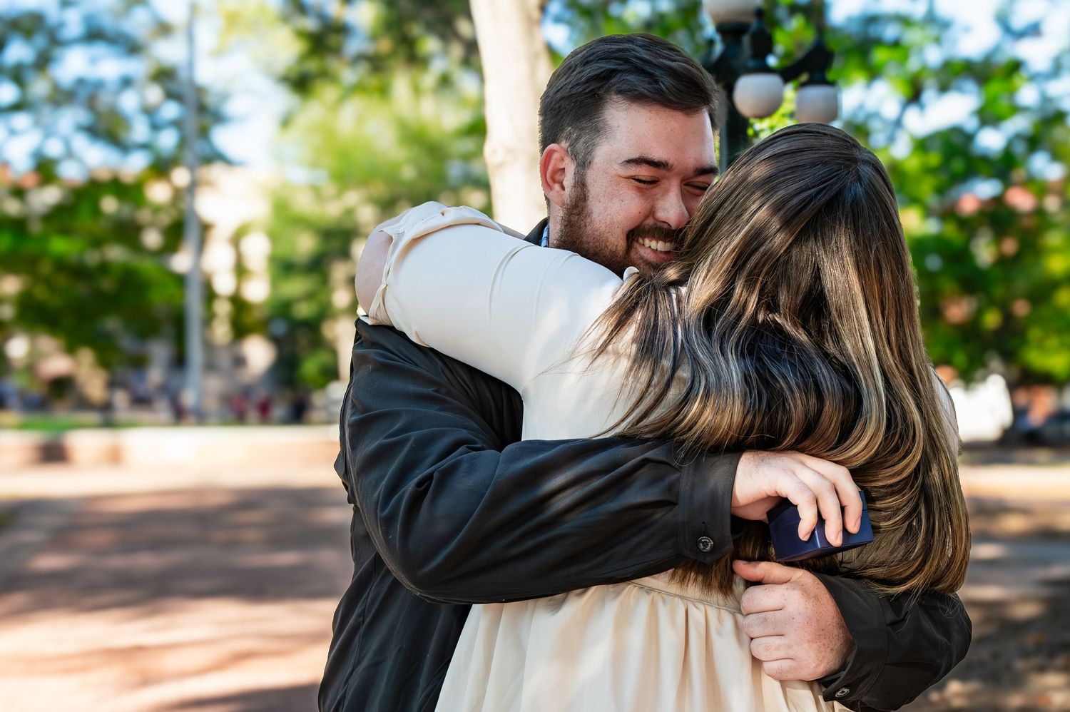 Two people share a warm embrace outdoors on a sunny day in a park with trees in the background.