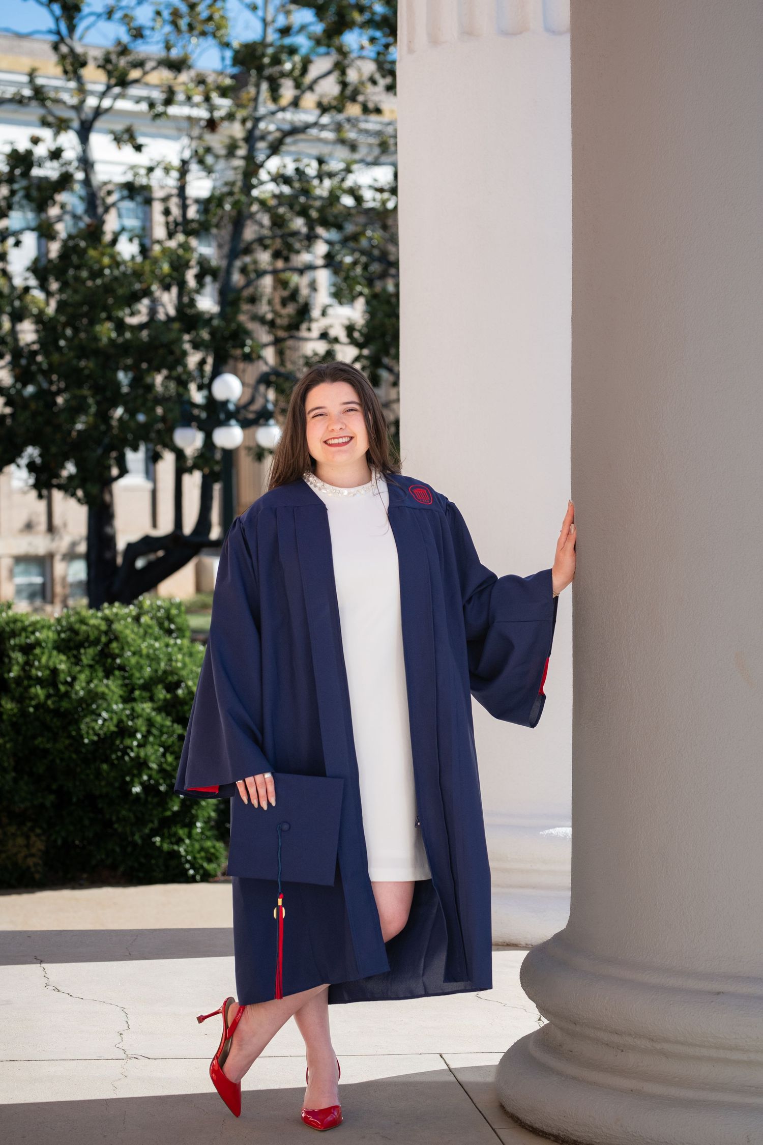 Smiling graduate in navy blue cap and gown with red heels posing by white columns outdoors.