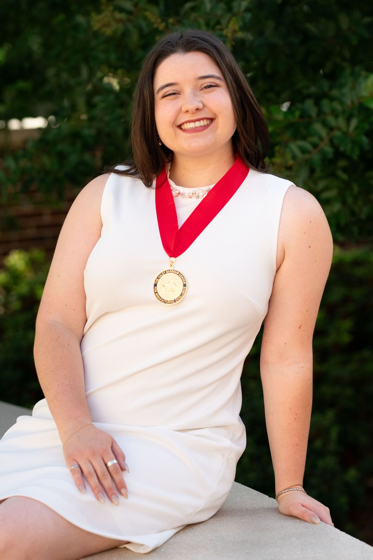 Smiling young woman in white dress wearing gold medal with red ribbon outdoors.