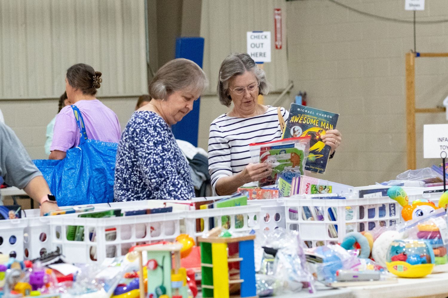 Two women browse books and toys at an indoor garage sale or community rummage sale event.