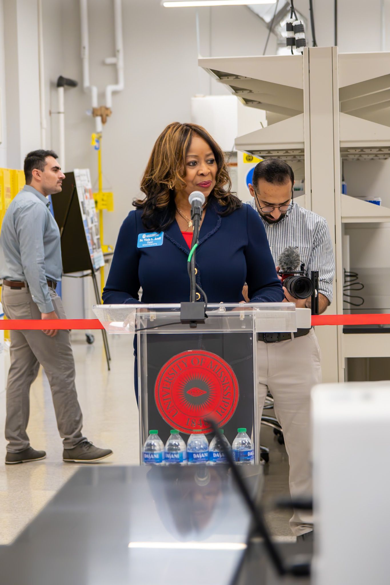 Woman speaking at podium with microphone at an indoor facility ribbon-cutting ceremony with staff in background.