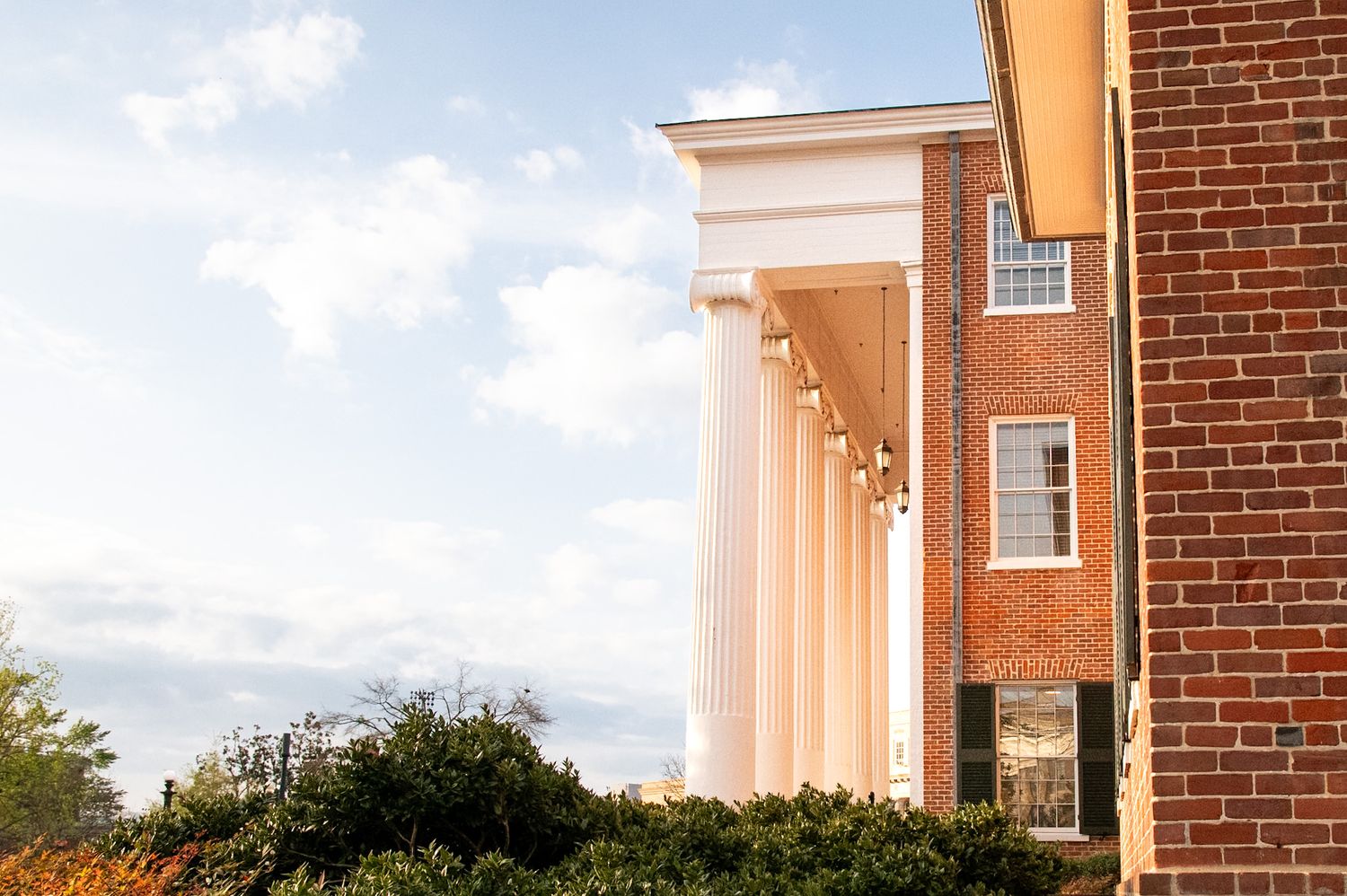 Classic brick building with white colonial columns against a blue sky, surrounded by lush green shrubs.