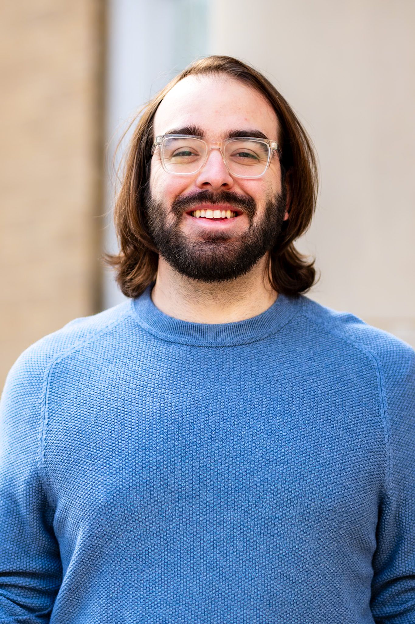A person with long dark hair, glasses, and a beard wearing a light blue sweater smiles warmly at the camera.