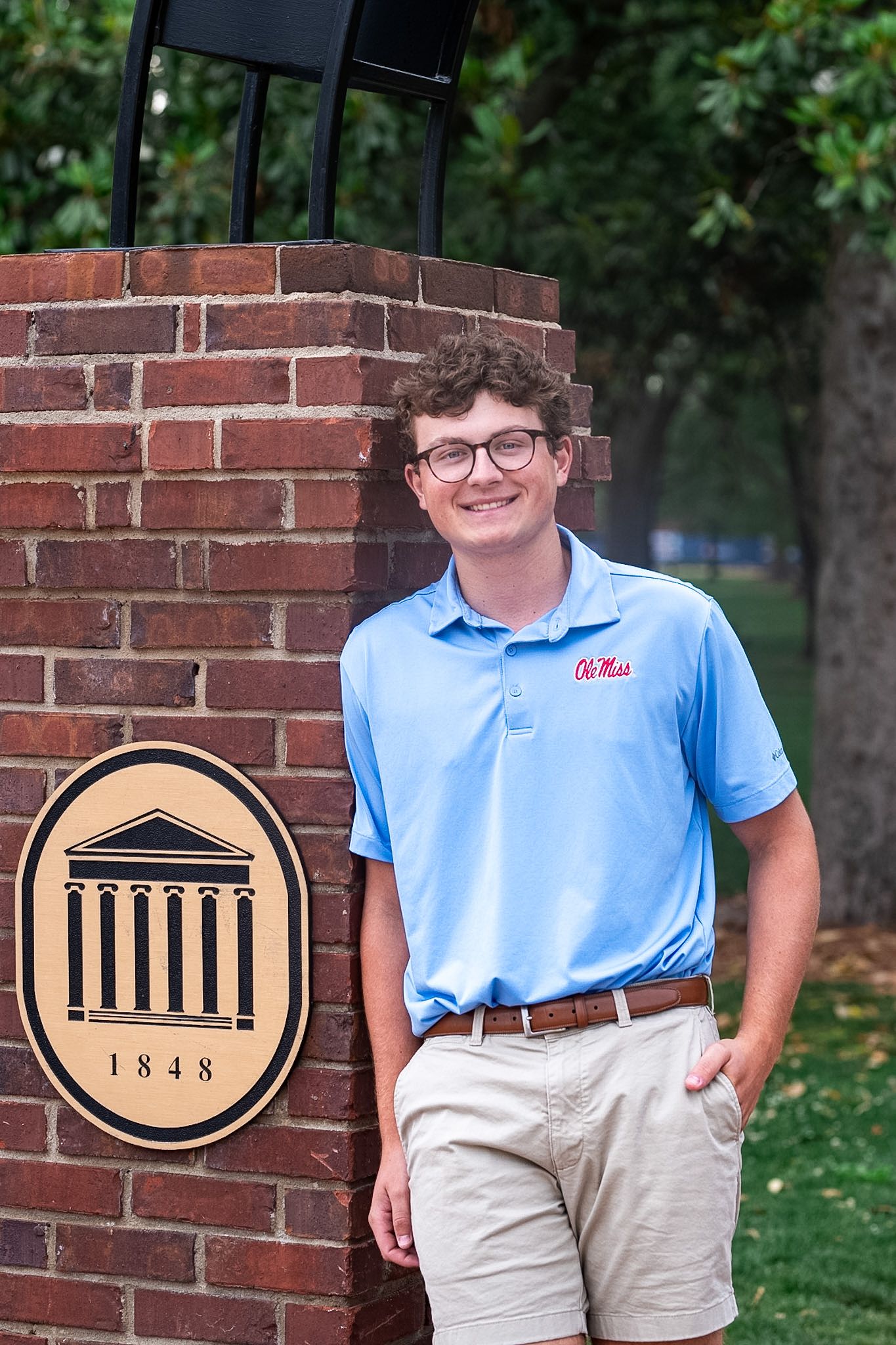 Young man in Ole Miss polo leaning against university brick entrance sign dated 1848 on sunny campus.