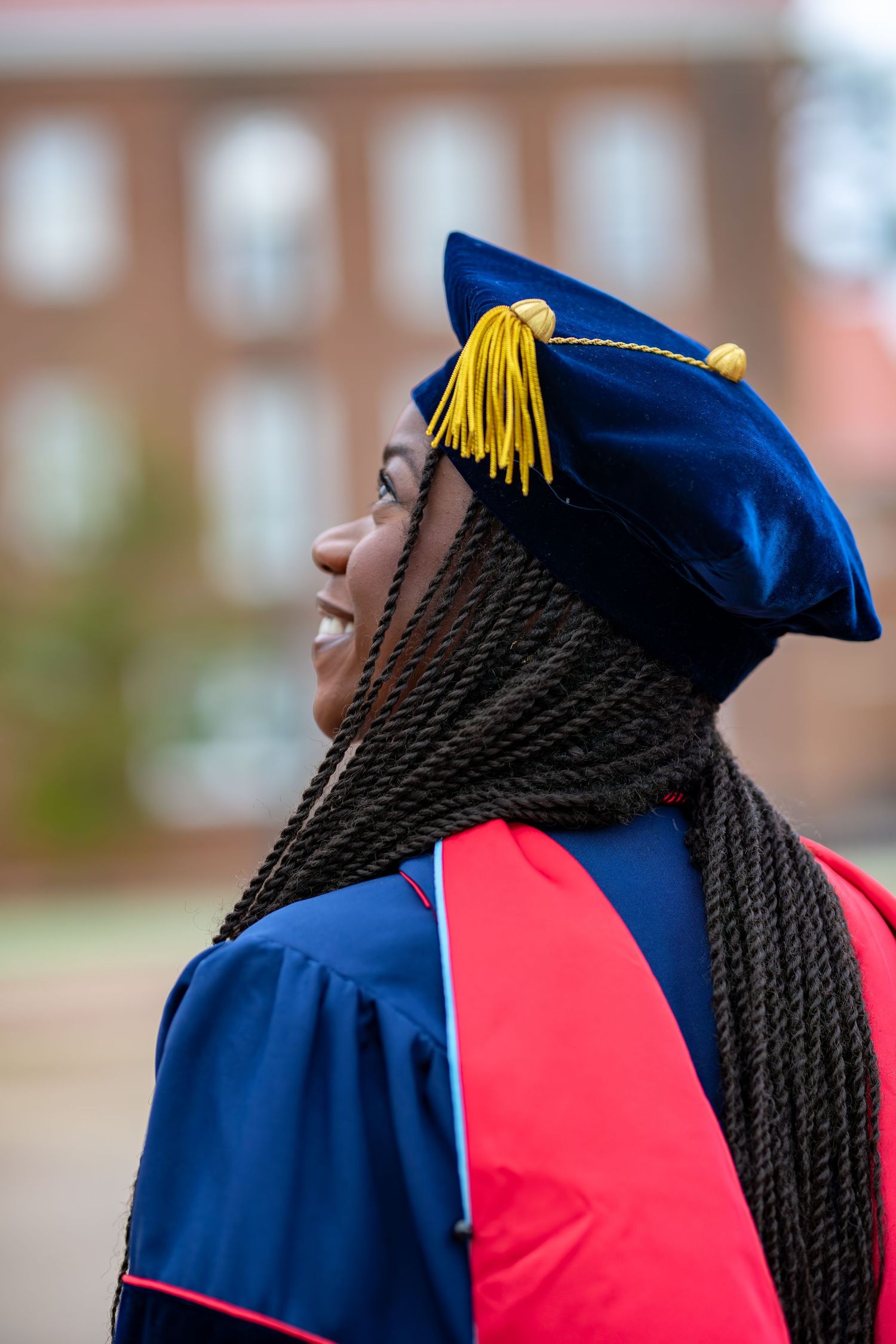 Smiling Black woman in blue doctoral graduation cap with gold tassel and red hood, looking up outdoors near brick building.
