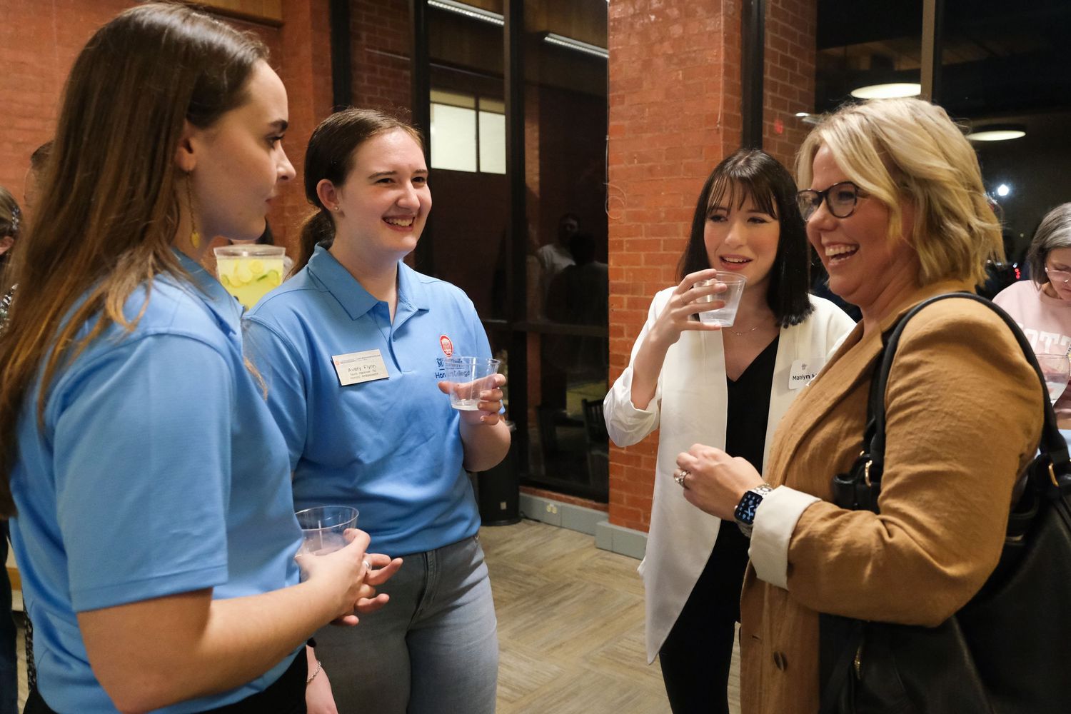 Four women socializing at a networking event, two wearing blue polo shirts with name tags, holding drinks.