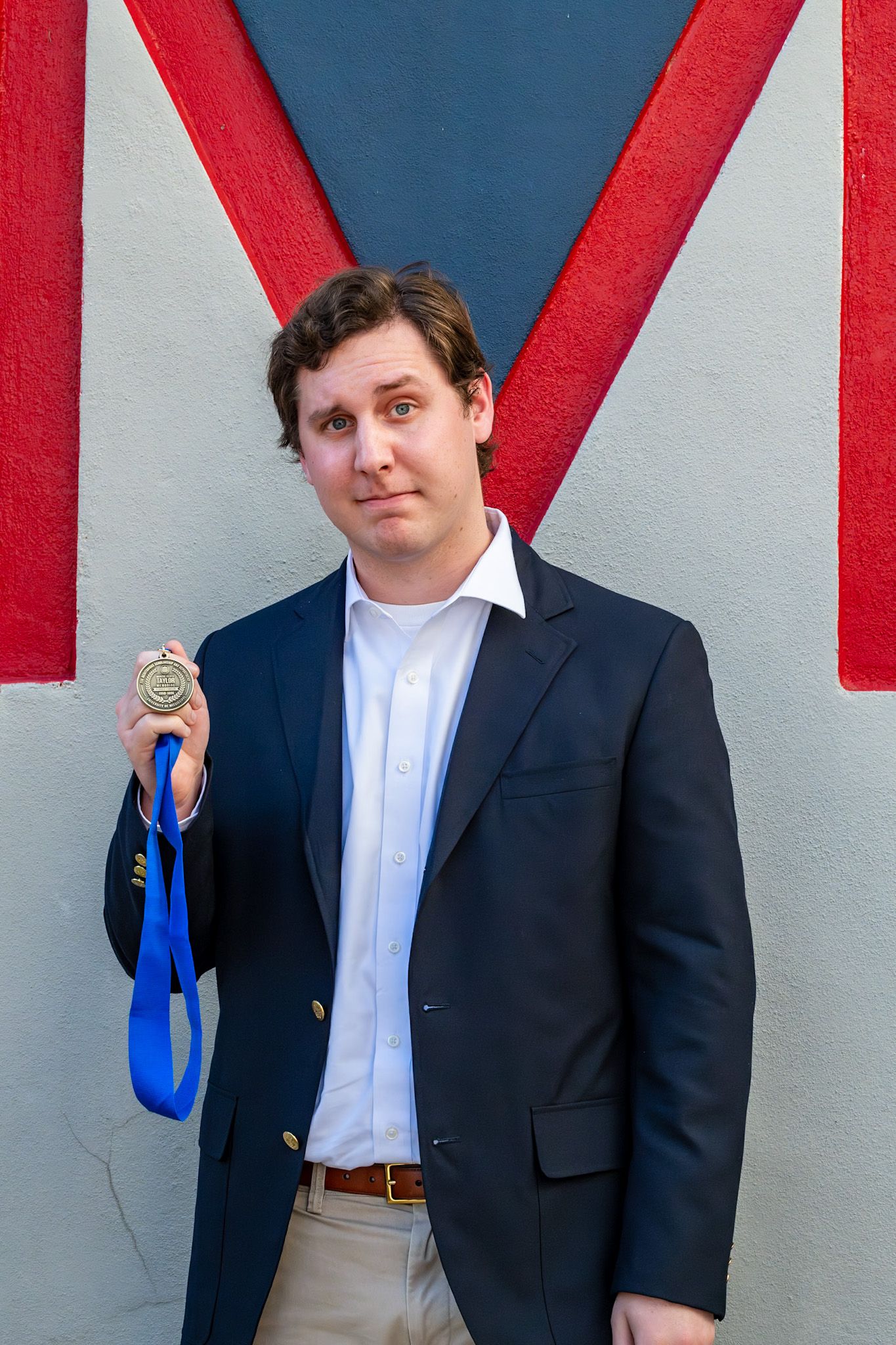 Young man in navy blazer proudly holding a gold medal with blue ribbon while posing against a decorative wall.