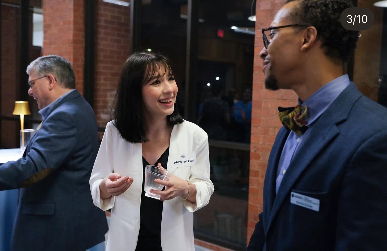 Two professionals in a white coat and suit converse at an indoor networking event with a third person in background.