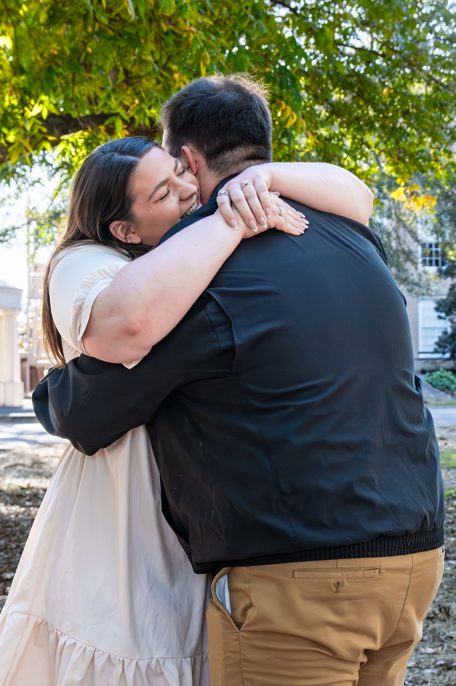 Two people share a warm embrace outdoors on a sunny day beneath green tree branches.