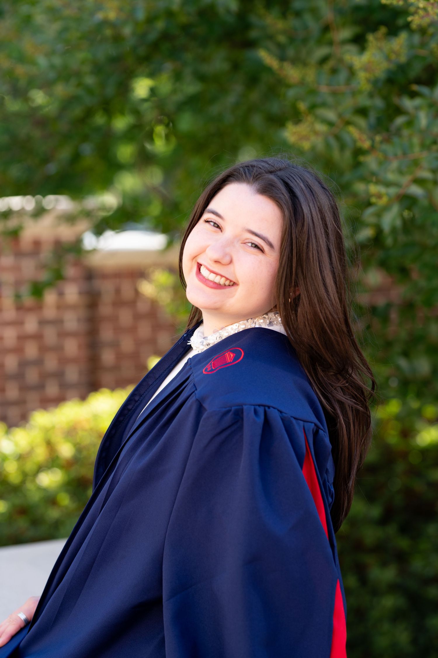 Smiling young woman in navy blue graduation gown posing outdoors with green trees and brick wall background.