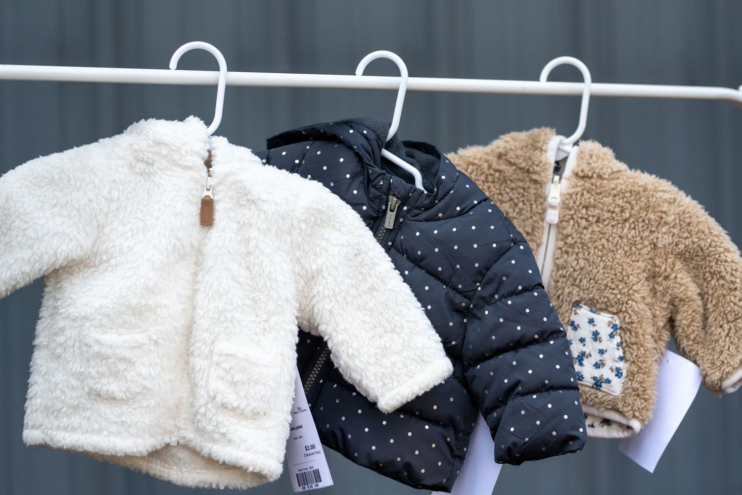 Three baby jackets on hangers: white fluffy, navy polka dot puffer, and tan teddy bear coat.
